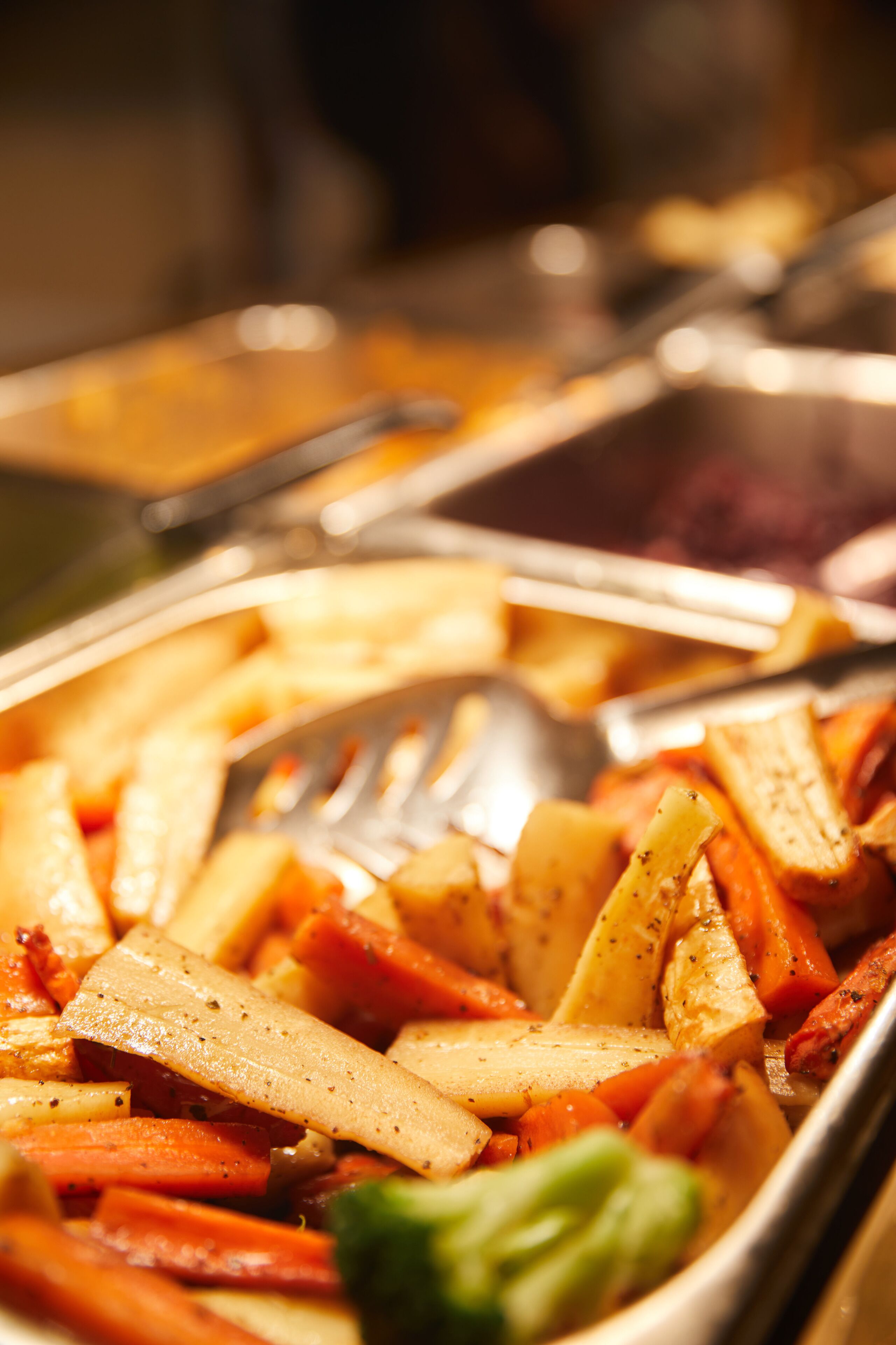 Primer plano de un bufé con diversos platos, que incluyen una mezcla de verduras y quizás algo de carne, servidos en una bandeja con un tenedor y una cuchara.