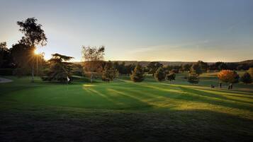 A golf course at sunset with a flagstick in the foreground, trees, and a clear sky.