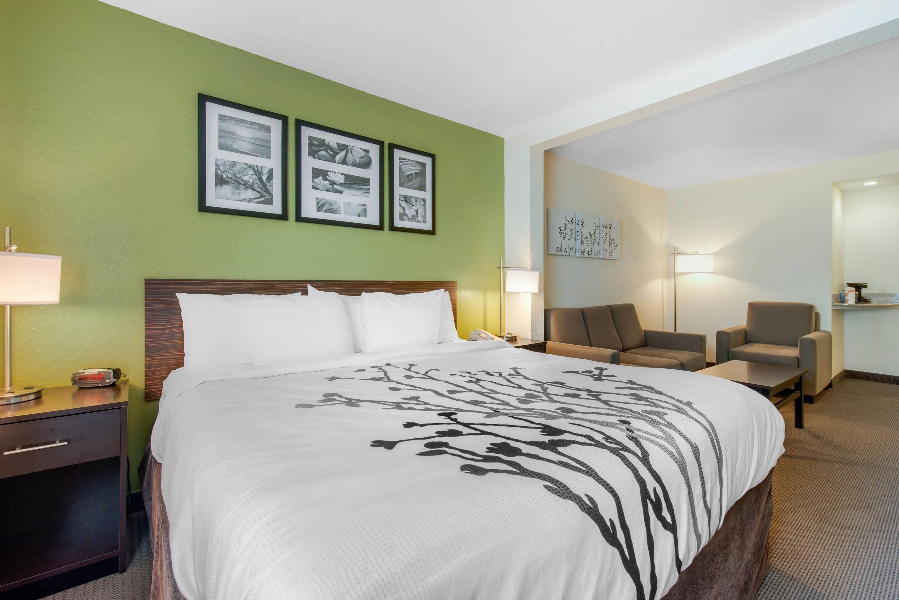 A hotel room with a large bed, two chairs, a desk, and a nightstand. There are framed pictures on the wall and a lamp on the desk.