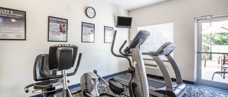 Un gimnasio con máquinas elípticas, cinta de correr y bicicletas estáticas. Hay carteles en la pared, un reloj y un televisor montado.