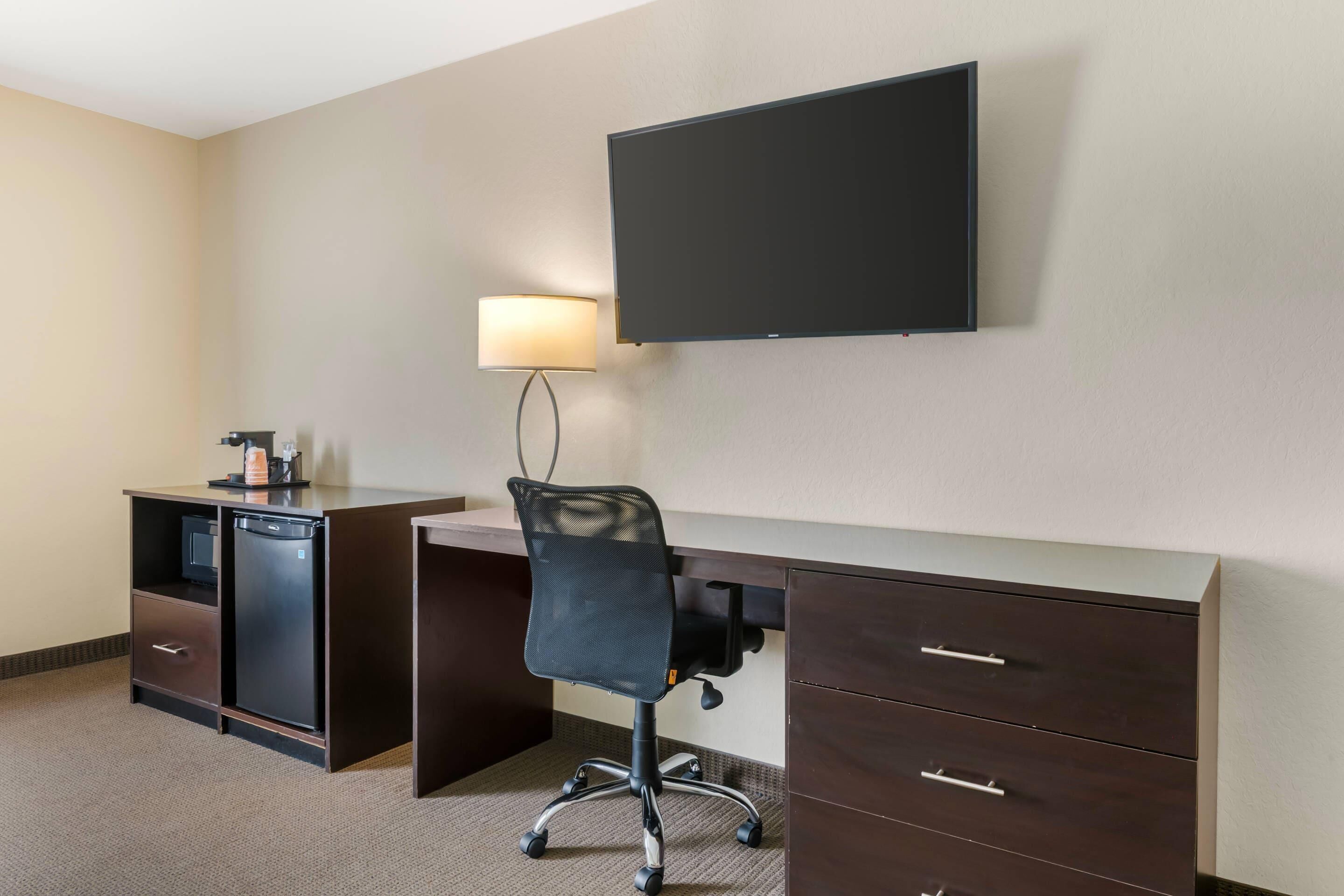A hotel room with a desk, chair, TV, lamp, and a minibar.