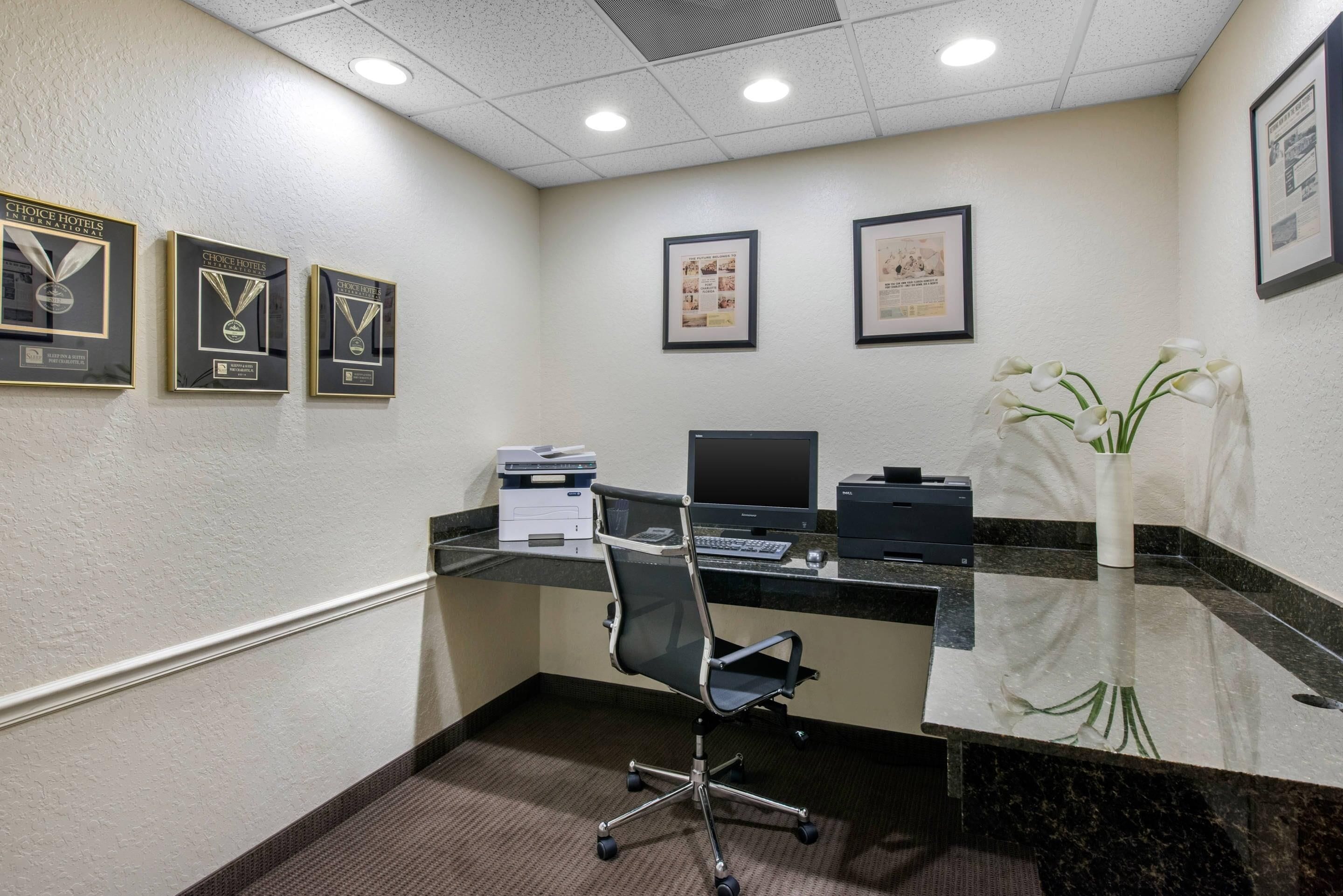 A conference room with a glass table, a black office chair, a desktop computer, and framed awards on the wall.