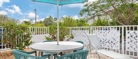 A round table with four teal chairs and a single lounge chair, under a striped blue and white umbrella, surrounded by a white fence with decorative elements.