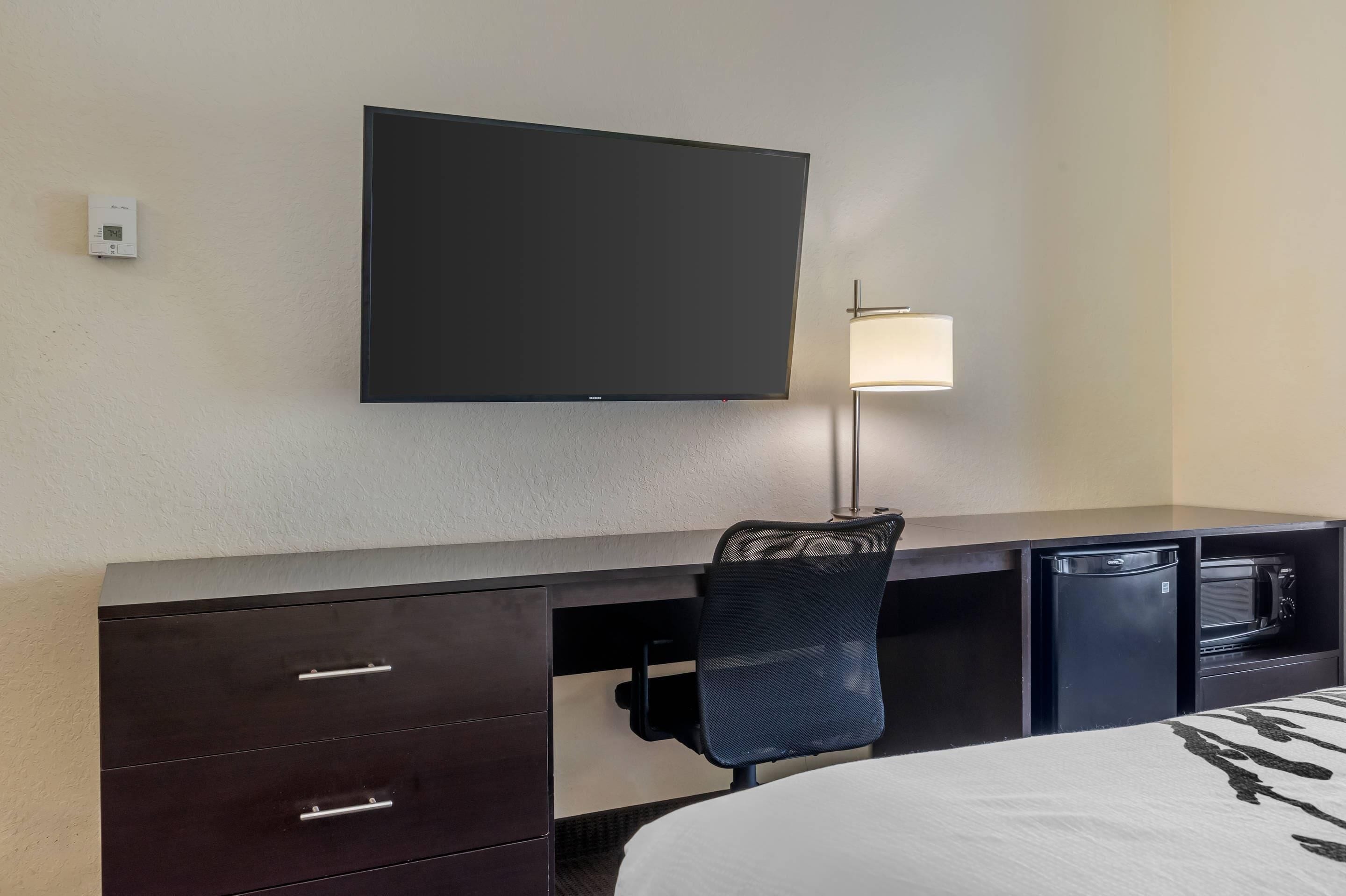 A hotel room with a flat-screen TV mounted on the wall, a desk with drawers, a chair, a small refrigerator, and a microwave.