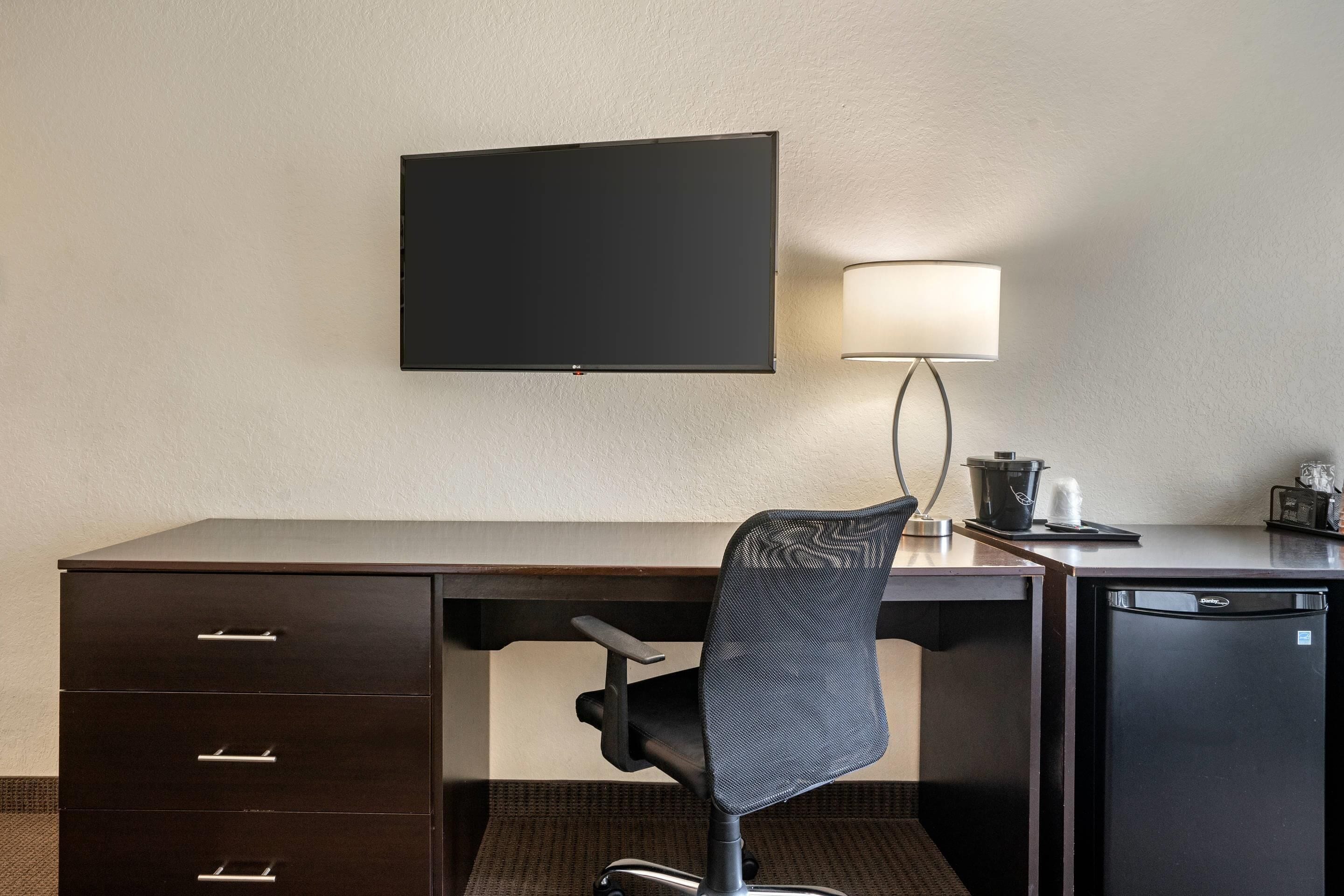 Une chambre d’hôtel avec un bureau en bois foncé, un fauteuil de bureau noir, un téléviseur à écran plat fixé au mur, une lampe de bureau, un petit réfrigérateur et une poubelle.