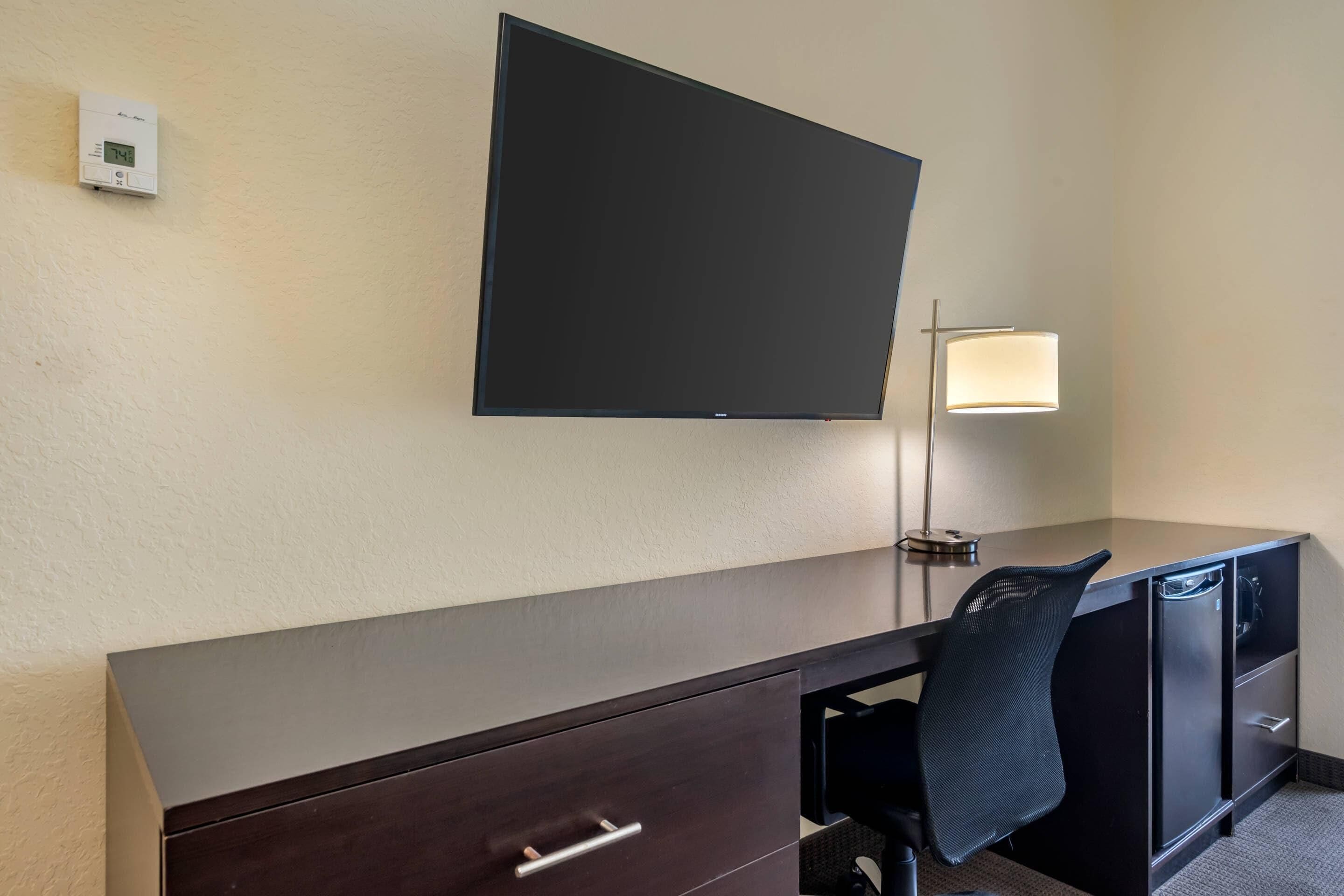 Habitación de hotel con televisor de pantalla plana montado en la pared, escritorio con silla, lámpara y termostato en la pared.