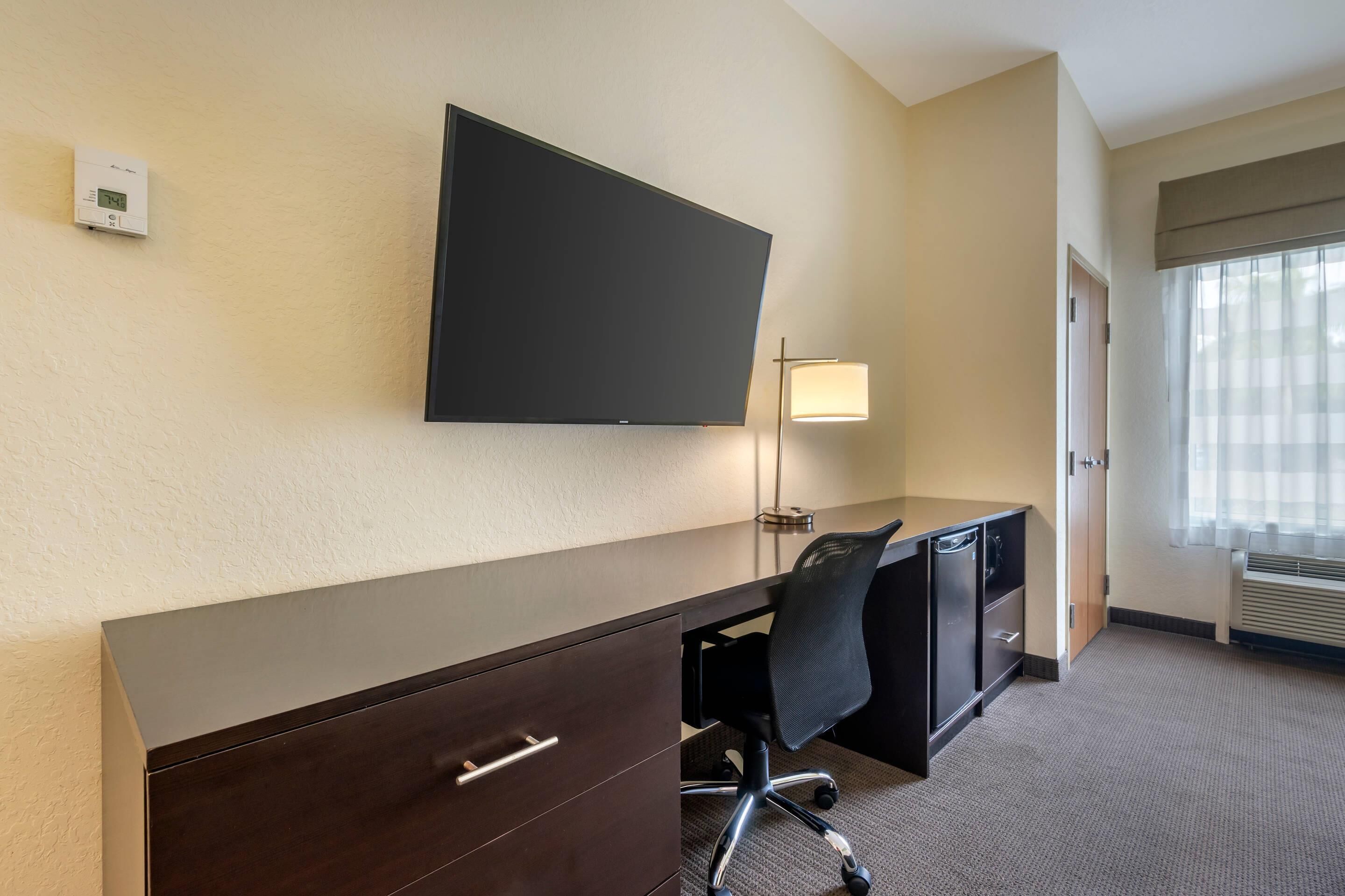 A hotel room with a flat-screen TV mounted on the wall, a desk with a chair, a lamp, and a window with blinds.