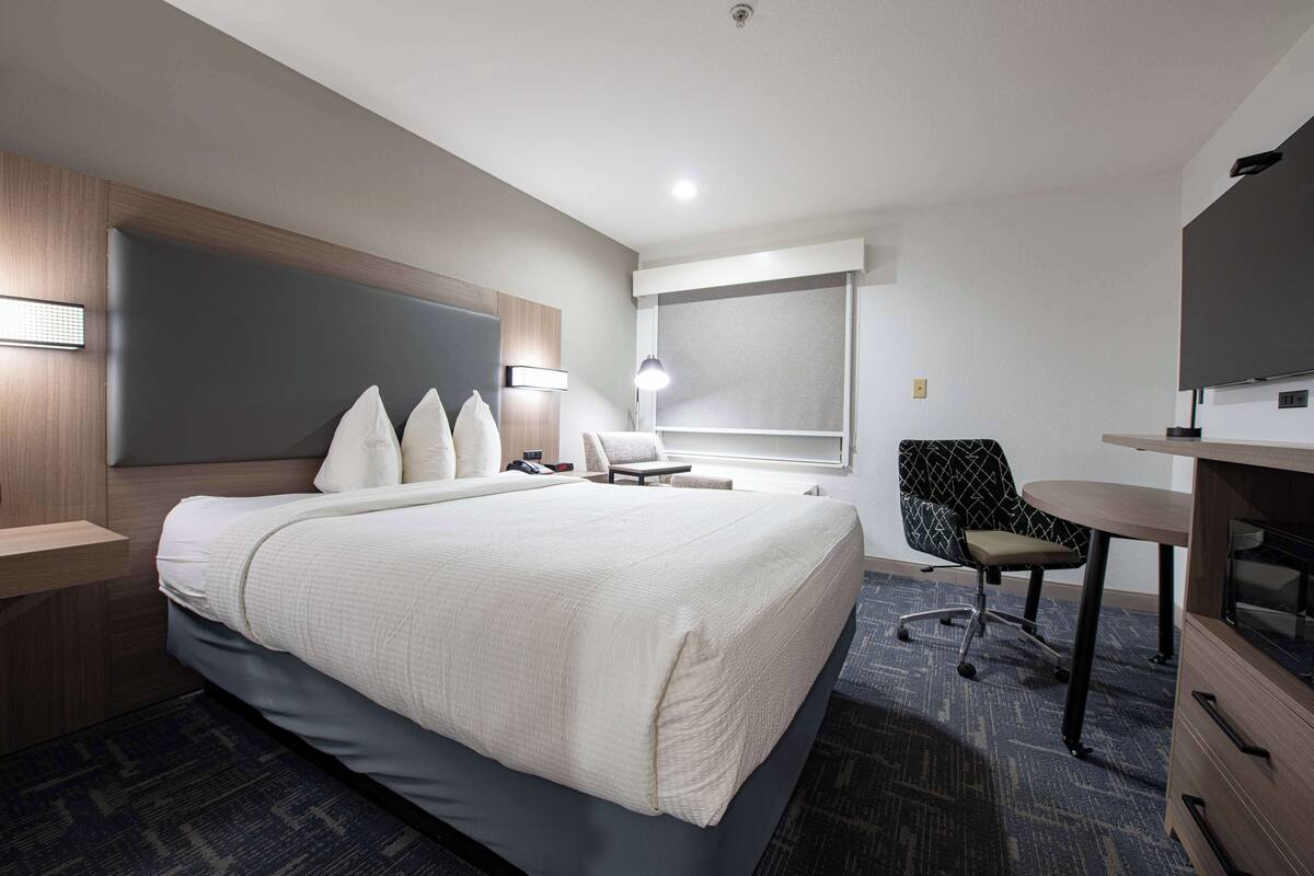 Una habitación de hotel moderna con una cama grande, un escritorio con televisor de pantalla plana, una silla y una lámpara de escritorio.