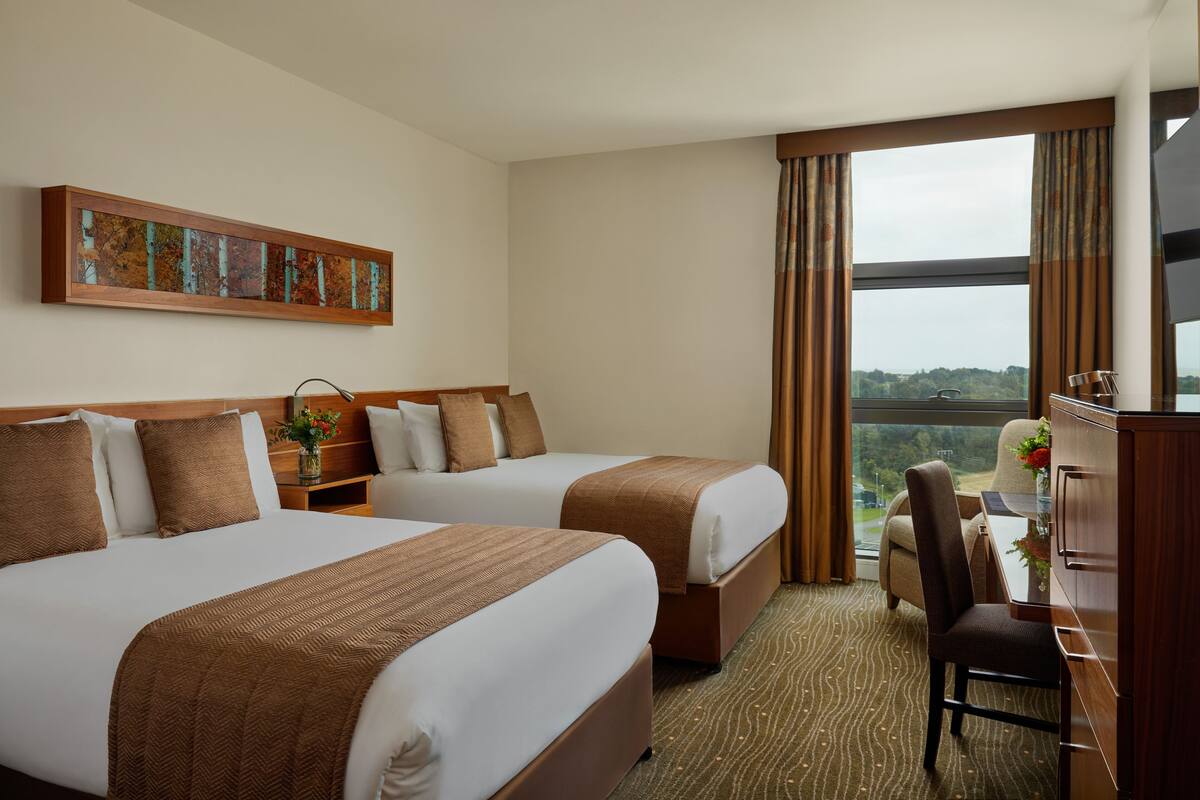 A hotel room with two beds, a desk, a chair, a TV, and a window with a view.