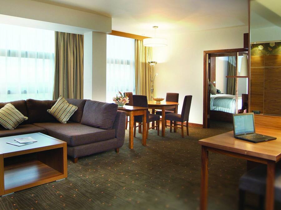 Habitación de hotel con sofá, una mesa de comedor con sillas, una computadora portátil sobre un escritorio y una cama con cabecera de madera.