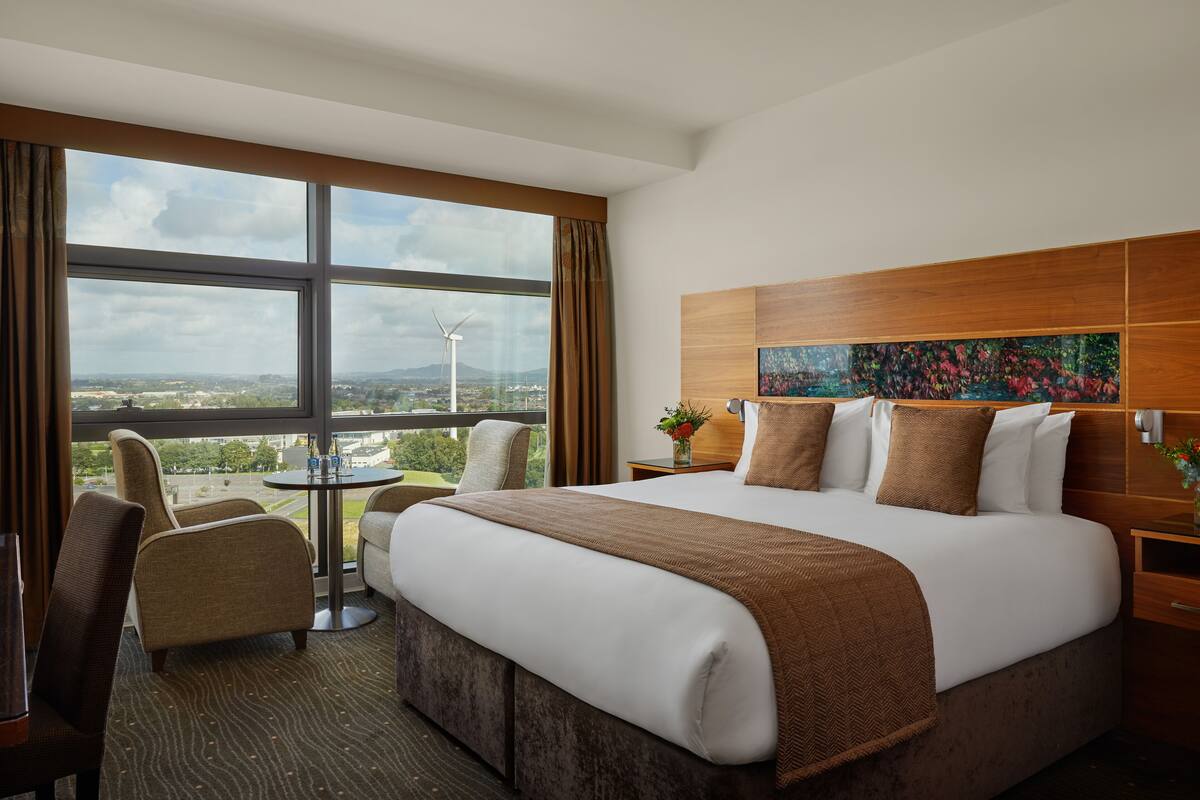 A hotel room with a large bed, a view of the cityscape, a round table, and a chair.