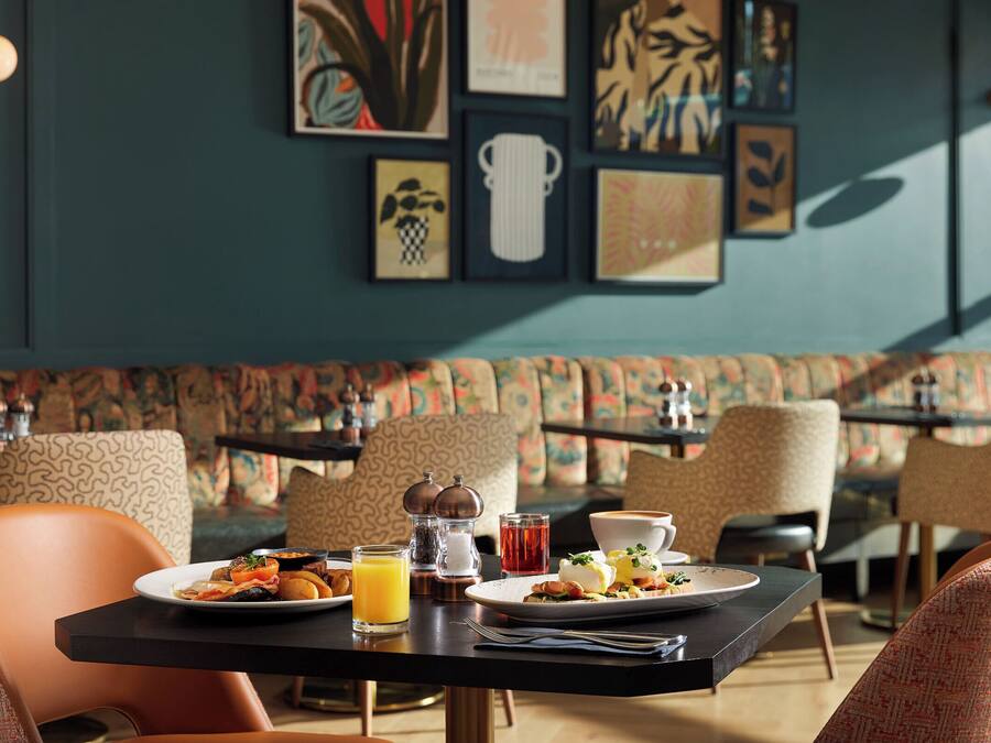 Un comedor con una mesa dispuesta para el desayuno, incluyendo platos de comida, bebidas y una taza de café. Se observan sillas con estampado y un banco acolchado al fondo.