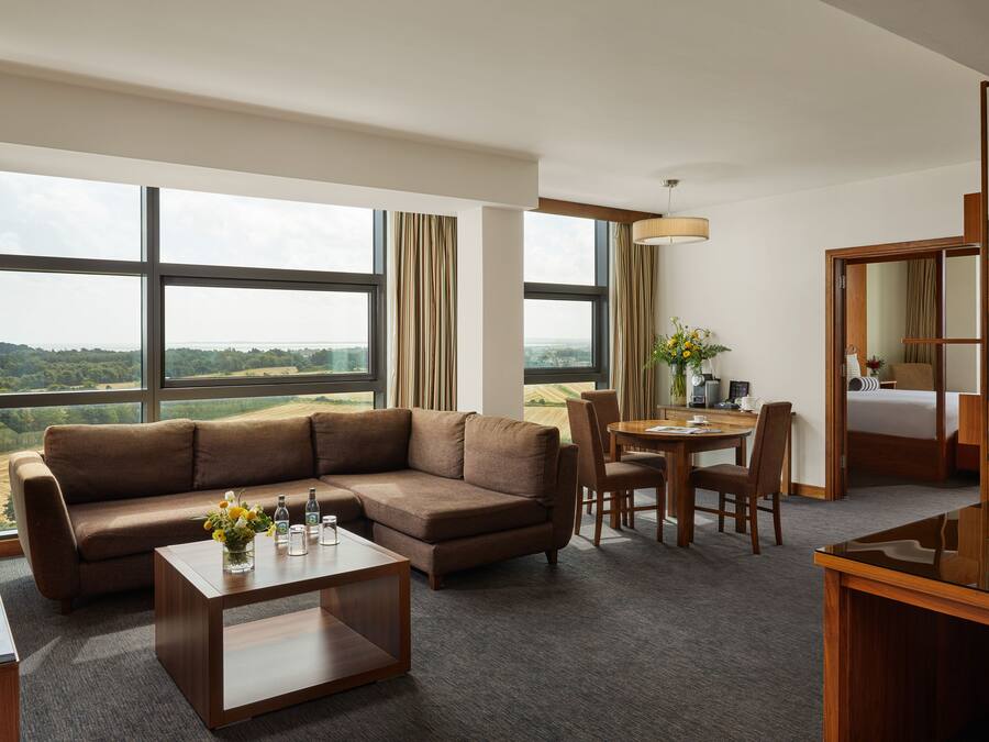 A modern hotel room with a large sectional sofa, a dining table with chairs, a small coffee table, and a large window offering a view of the countryside.