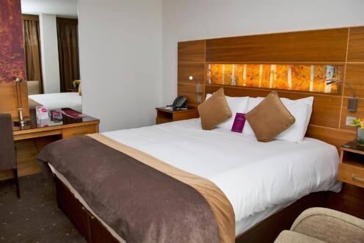 A hotel room with a large bed, a wooden headboard, a bedside table with a lamp, and a chair.
