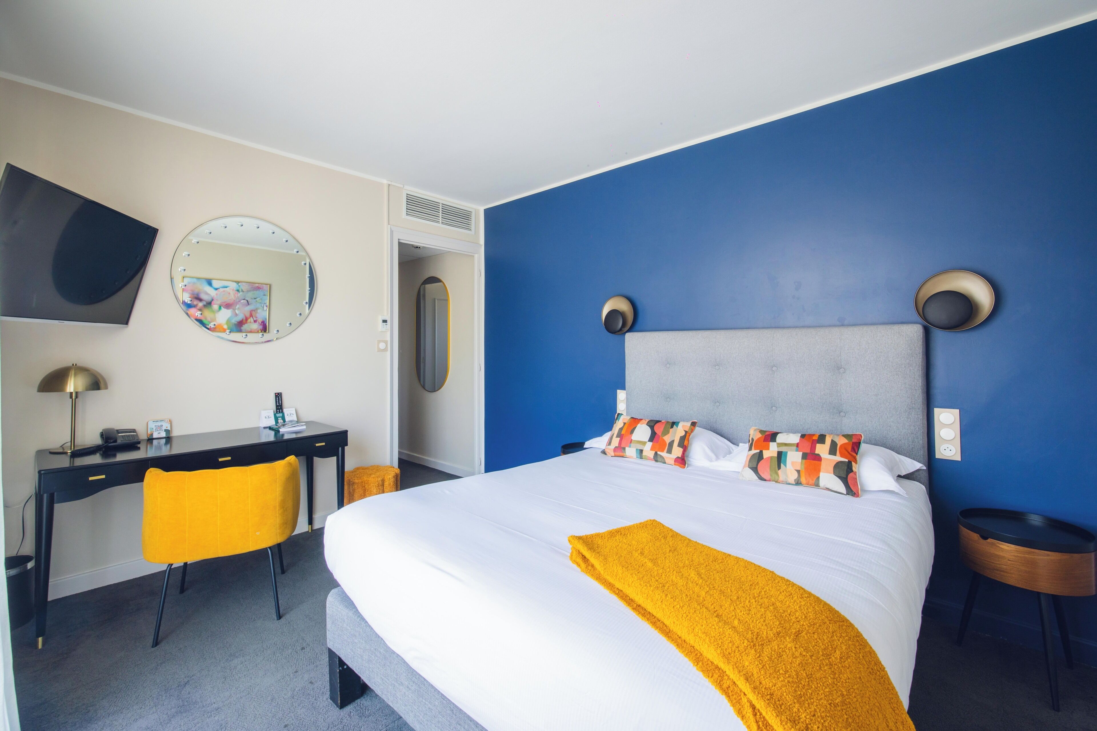 A hotel room with a bed, a desk, a chair, a mirror, and a round table.
