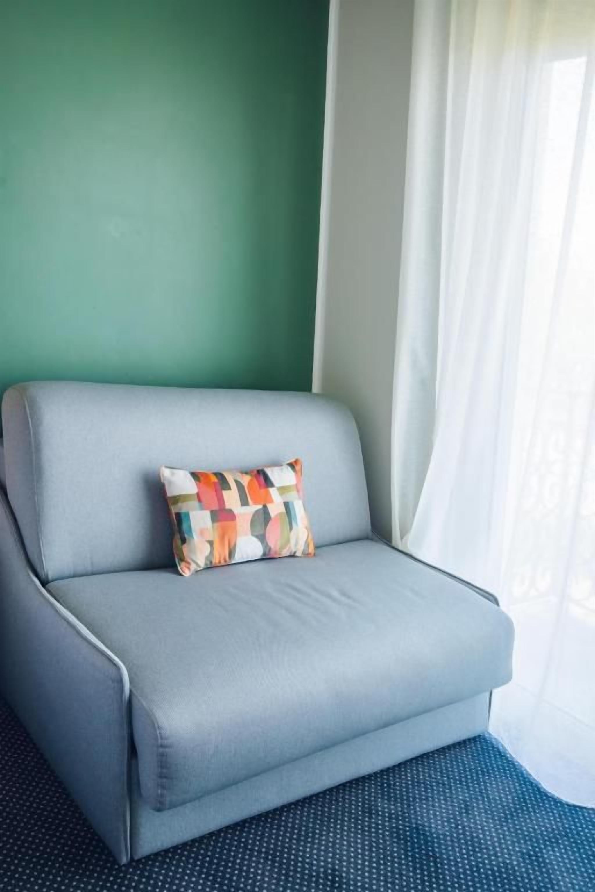 A light grey sofa with a colorful geometric pillow against a teal wall, positioned next to a window with white curtains.