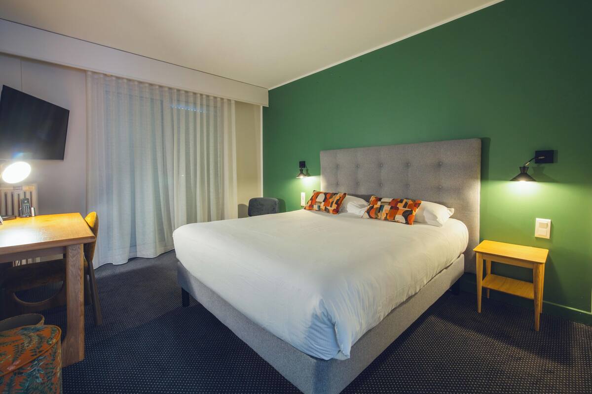 Habitación de hotel con una cama grande, un escritorio, una silla, un televisor y una ventana con cortinas.