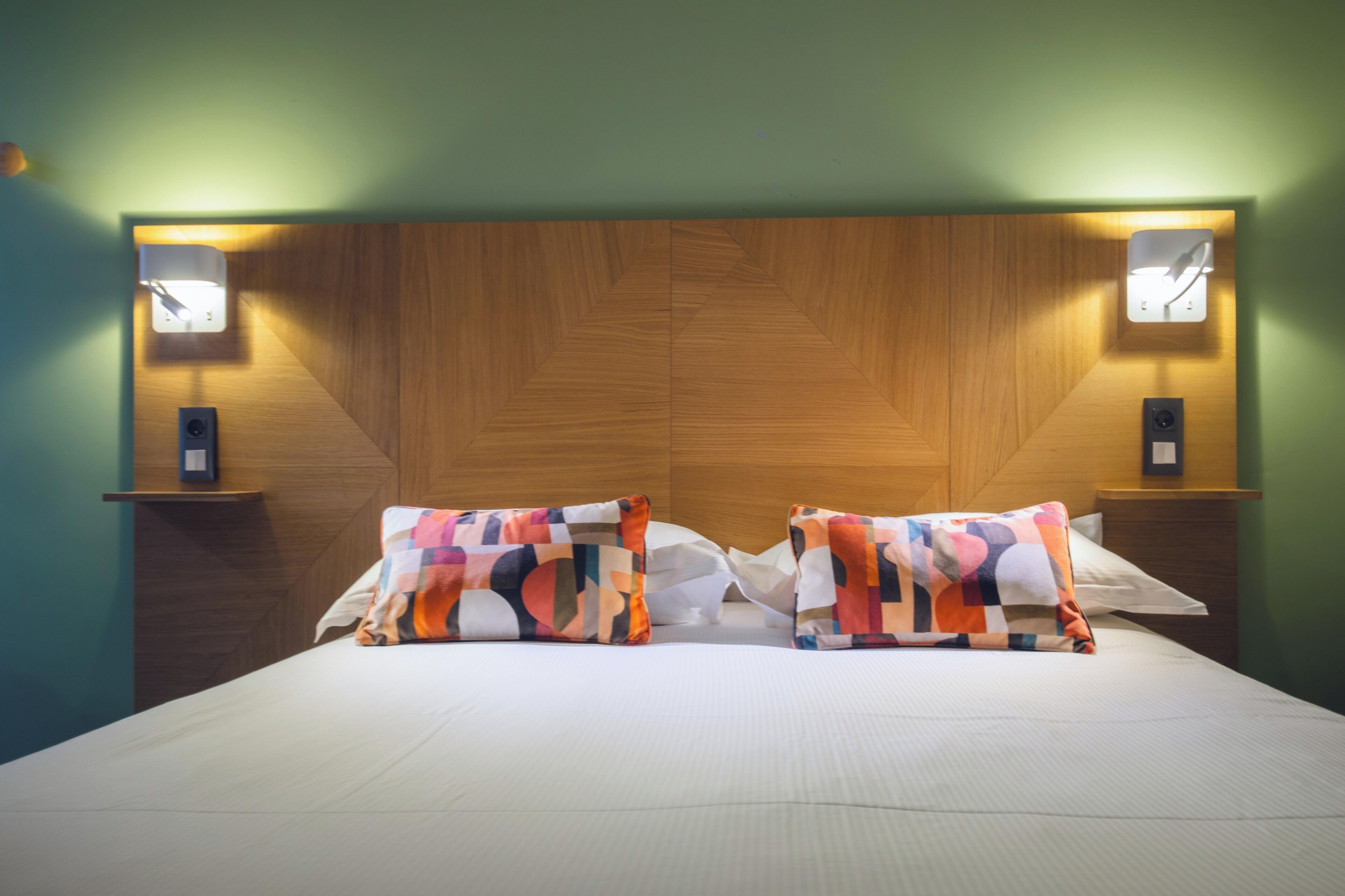 A hotel room with a wooden headboard, two bedside lamps, and a bed with patterned pillows.