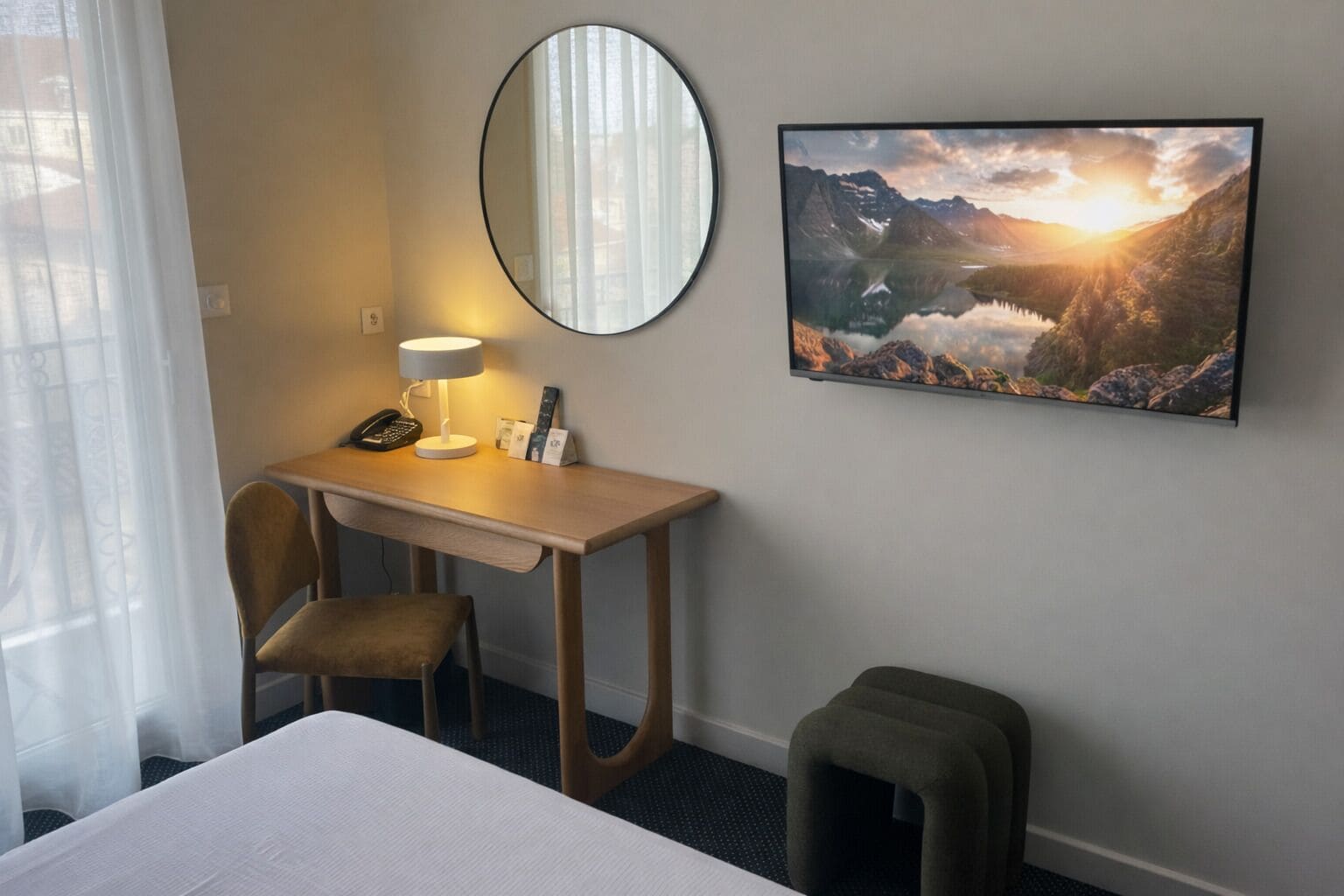 A hotel room with a bed, a desk with a lamp, a chair, a small stool, a mirror, and a framed picture of a mountain landscape.