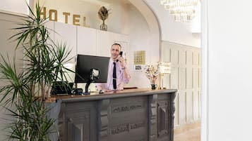 Un comptoir d’accueil d’hôtel avec un écran d’ordinateur, un téléphone et un vase contenant des fleurs séchées.
