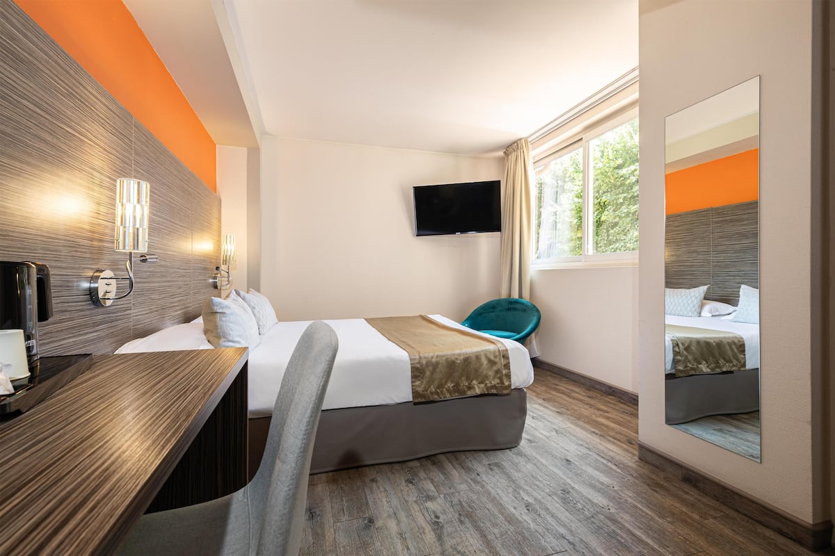 Habitació d'hotel amb llit, escriptori, televisió i finestra amb vista a arbres.