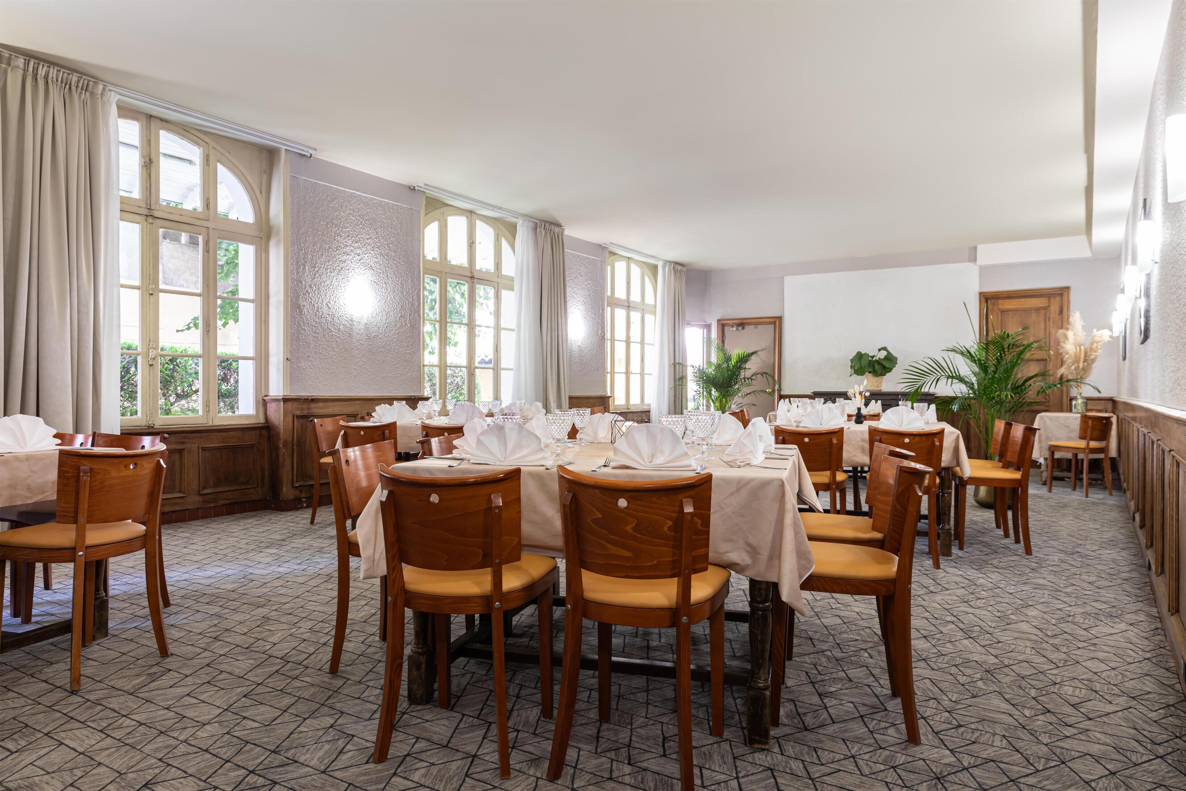 A dining area with multiple tables set for service, wooden chairs, large windows with curtains, and a tiled floor.