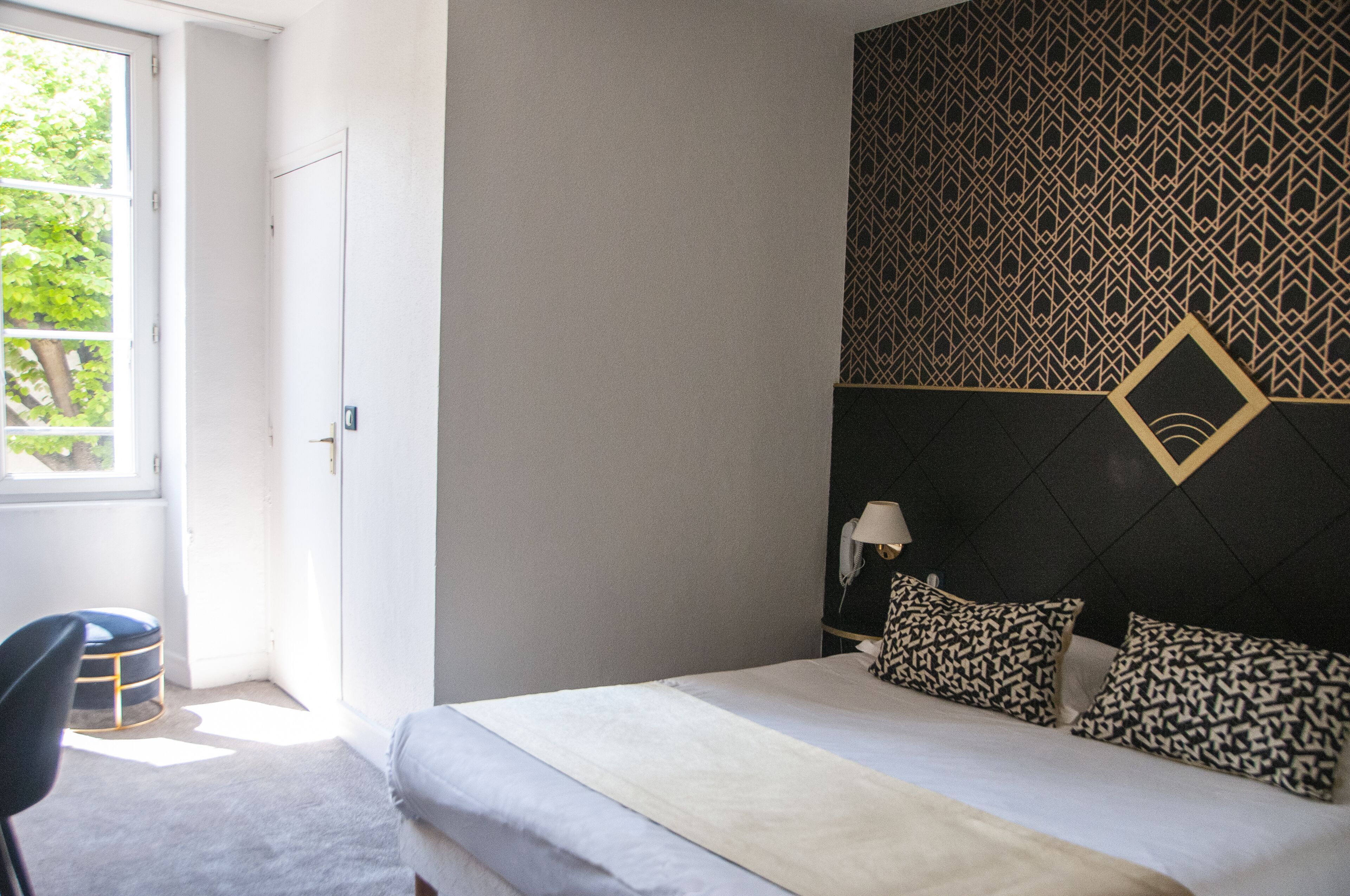 A bedroom with a bed, a patterned headboard, a nightstand with a lamp, and a window with a view of greenery.