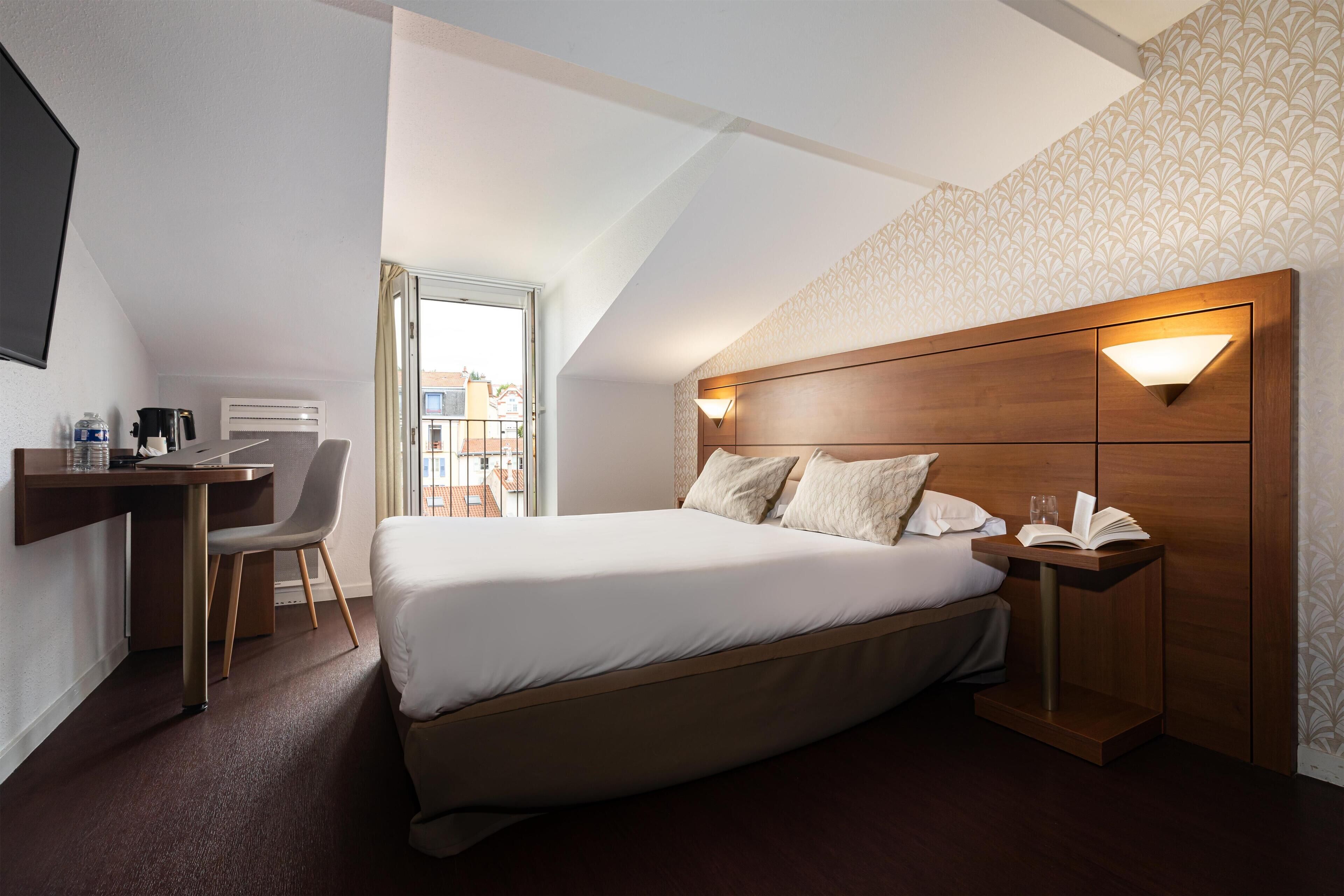 A modern hotel room with a large bed, a desk, and a chair. There is a window providing a view of the outside.