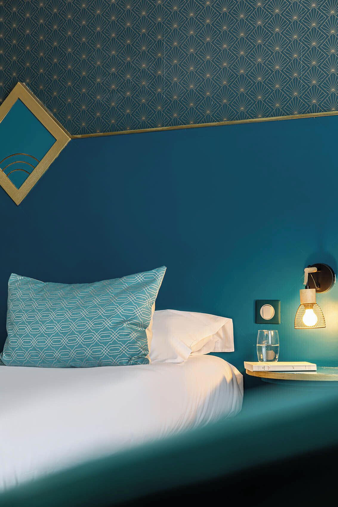 A neatly made bed with a teal headboard and a patterned pillow, a bedside table with a lamp, and a framed picture on the wall.