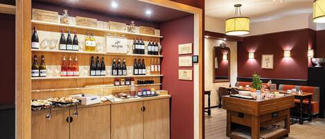Una bodega con estantes de madera que exhiben diversas botellas de vino y copas, un mostrador de madera con pasteles, y un comedor con mesa y sillas.