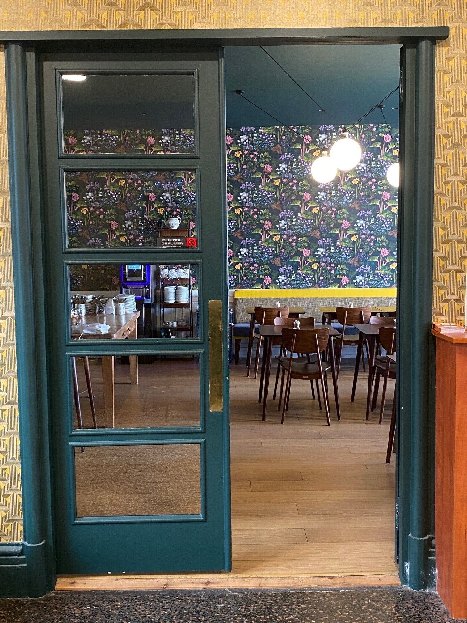L’intérieur d’un restaurant, avec une porte de couleur bleu-vert foncé, un papier peint à motifs floraux et un coin bar doté de tabourets et de tables.
