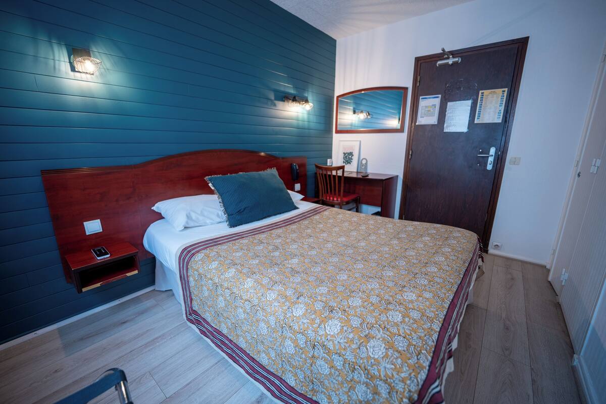 A bedroom with a wooden headboard, a bed with a patterned bedspread, a nightstand with a phone, a desk with a chair, and a door with a mirror and papers.