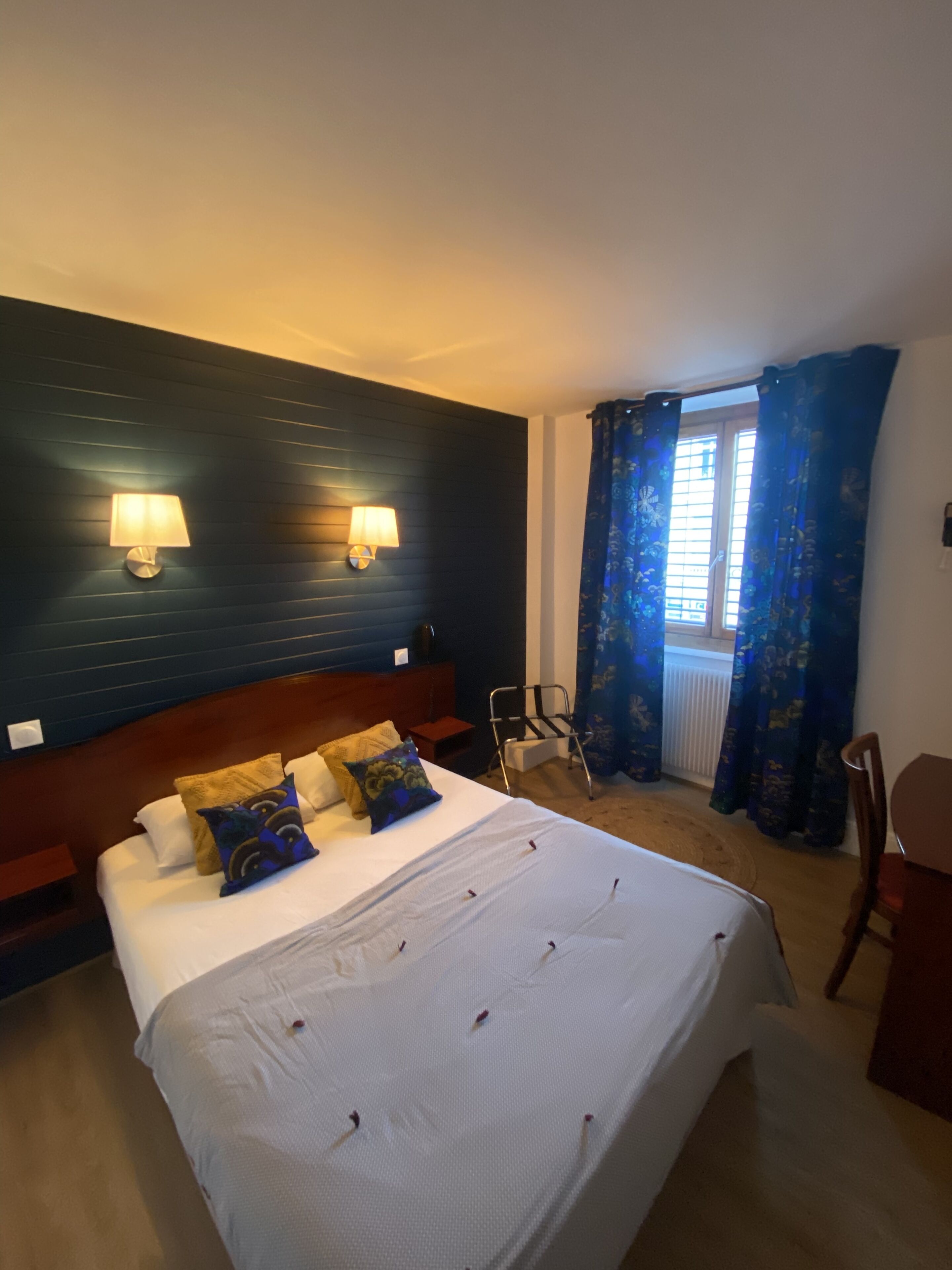 A hotel room with a large bed, two wall-mounted lamps, a window with blue curtains, and a chair.