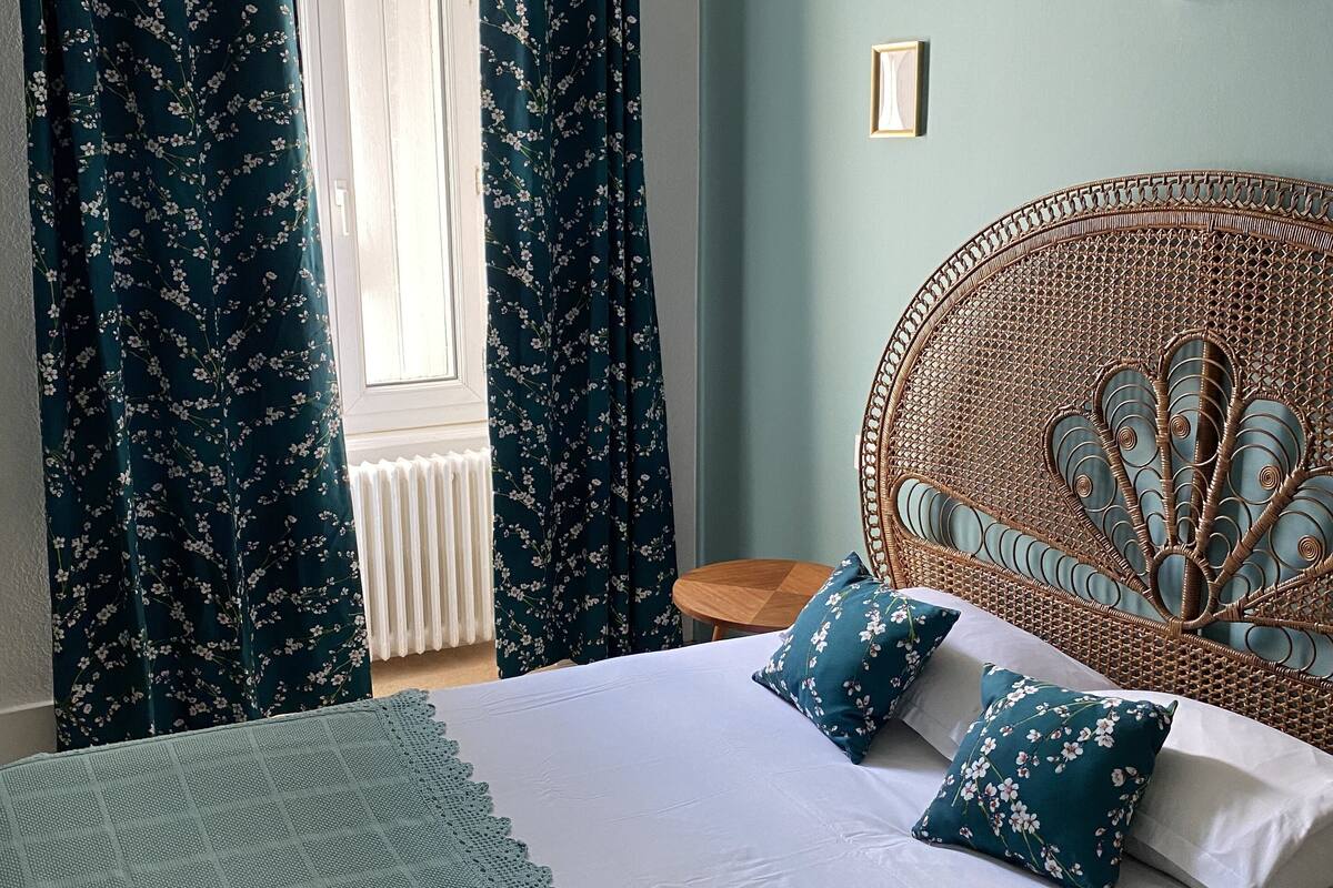 A bedroom with a bed, floral curtains, a rattan headboard, and a small wooden side table.