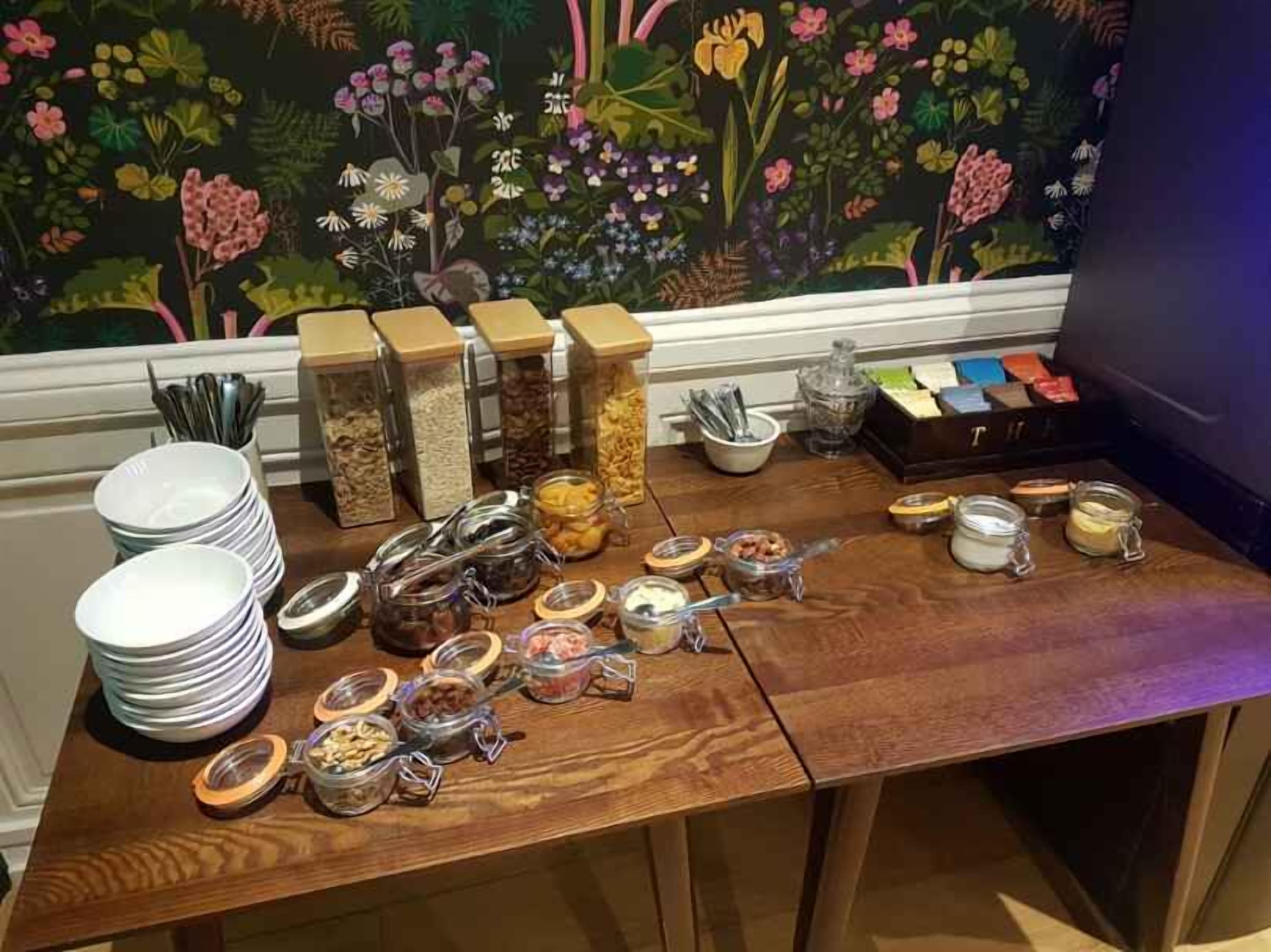 Una mesa de buffet con diversos alimentos y condimentos, incluyendo tarros de frutos secos y salsas, con un mural floral de fondo.