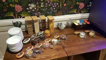 Una mesa de buffet con diversos alimentos y condimentos, incluyendo tarros de frutos secos y salsas, con un mural floral de fondo.