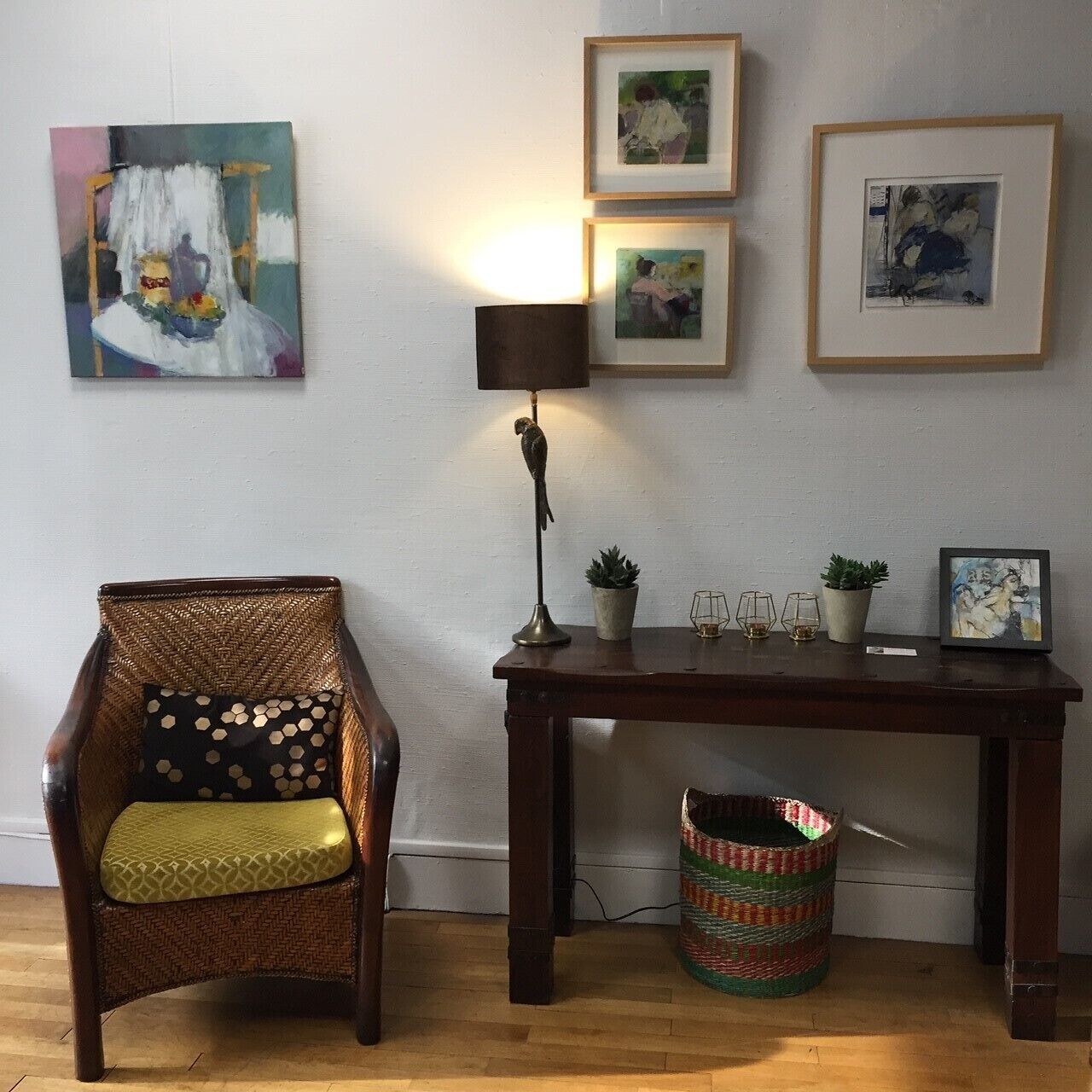 Une pièce comprenant un fauteuil en osier, une table en bois ornée de plantes en pot, des œuvres d’art encadrées accrochées au mur et une lampe.