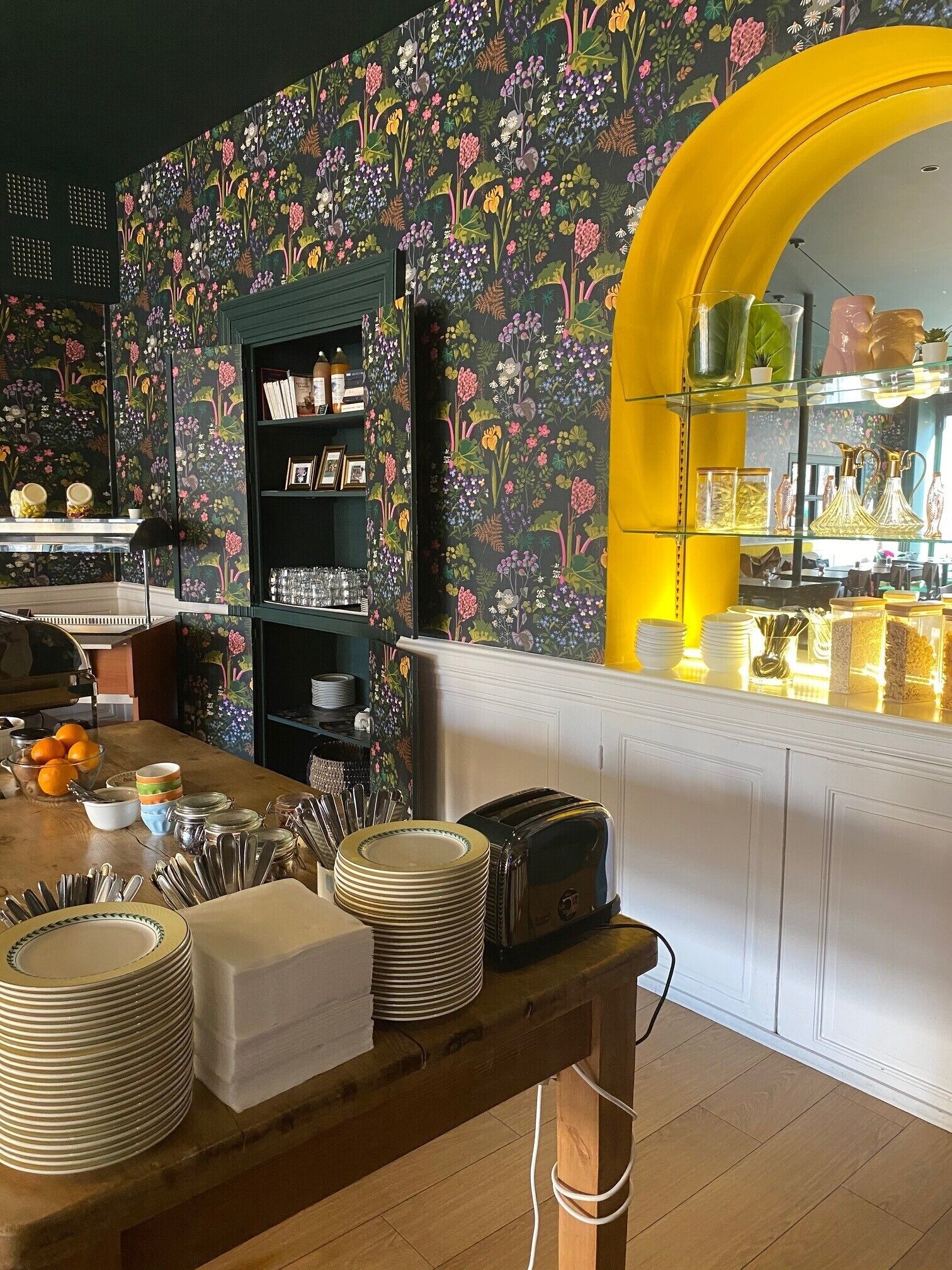 A restaurant interior with a floral wallpaper, a wooden table with stacked plates, a vintage toaster, and a yellow archway leading to the bar area.