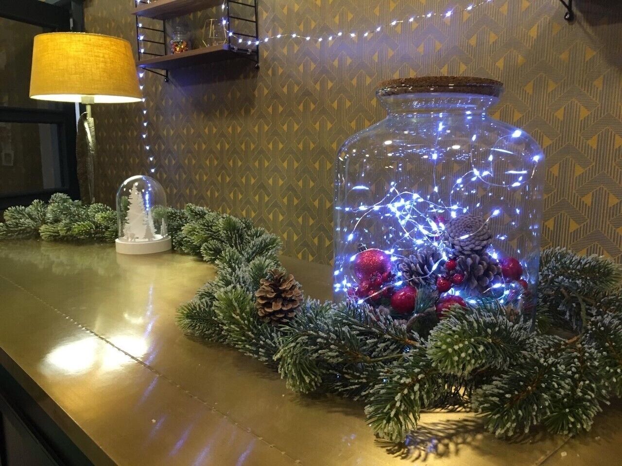 A decorative glass jar filled with pine cones and red ornaments, surrounded by a wreath on a polished surface.
