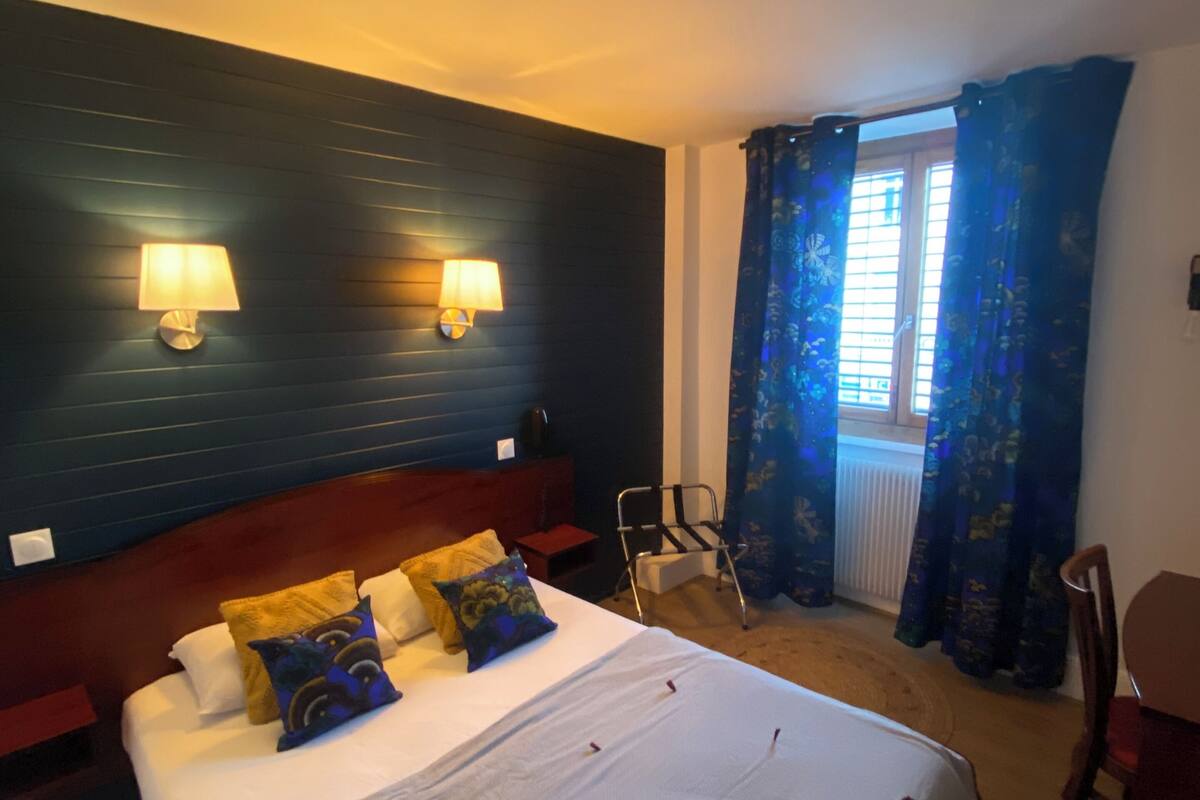 A hotel room with a large bed, two wall-mounted lamps, a window with blue curtains, and a chair.
