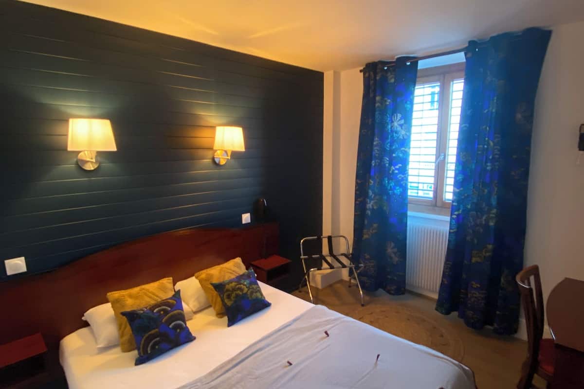 Habitación de hotel con una cama grande, dos lámparas de pared, un ventanal con cortinas azules y una silla.