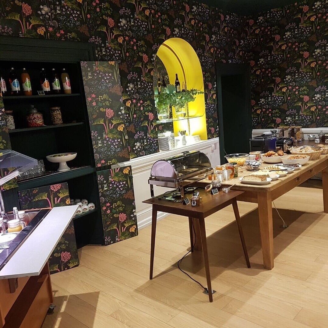Una habitación con una larga mesa de madera dispuesta para el desayuno, rodeada de un papel tapiz floral, una estantería y una pequeña planta.