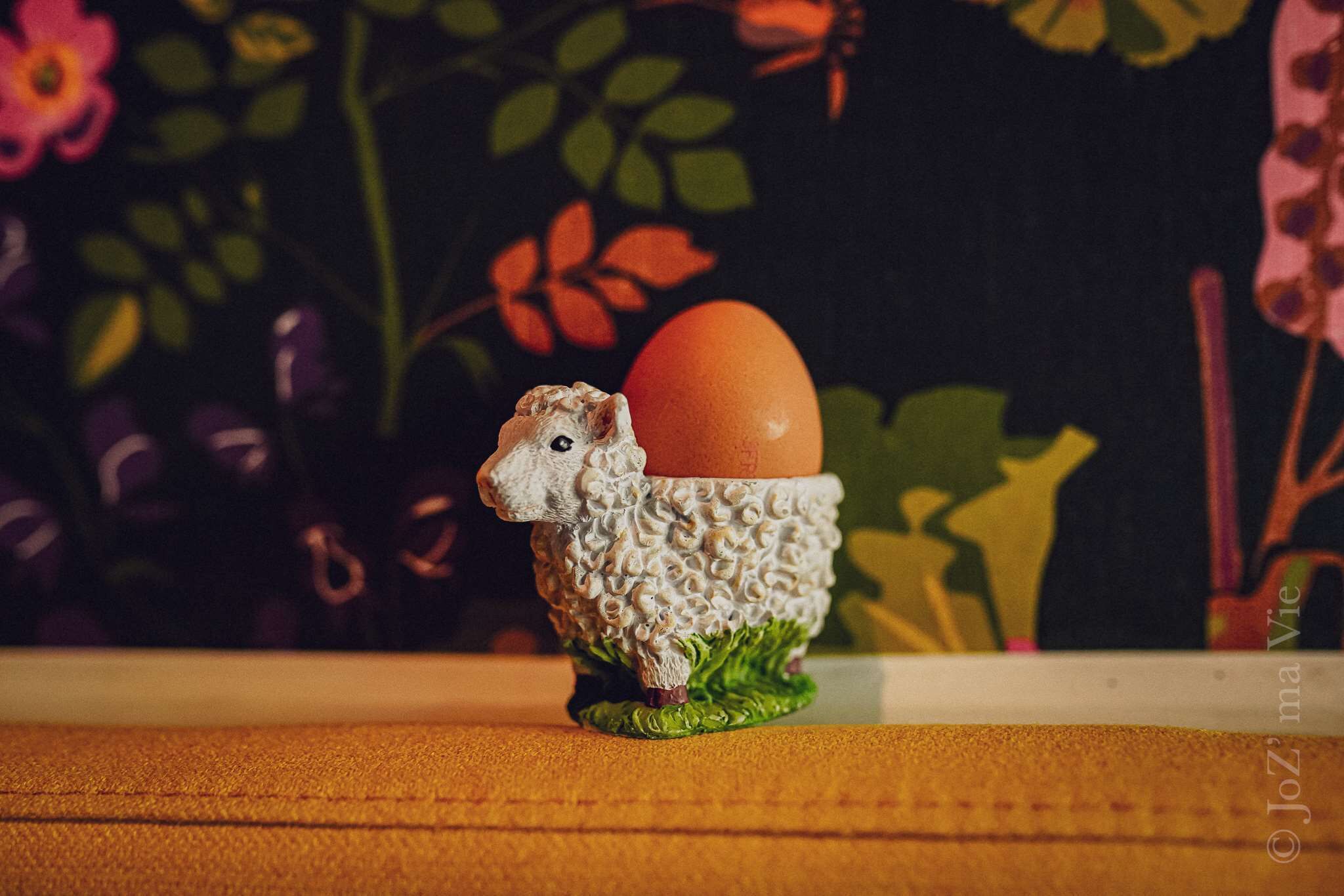A ceramic sheep figurine holding an egg on a table with a floral background.