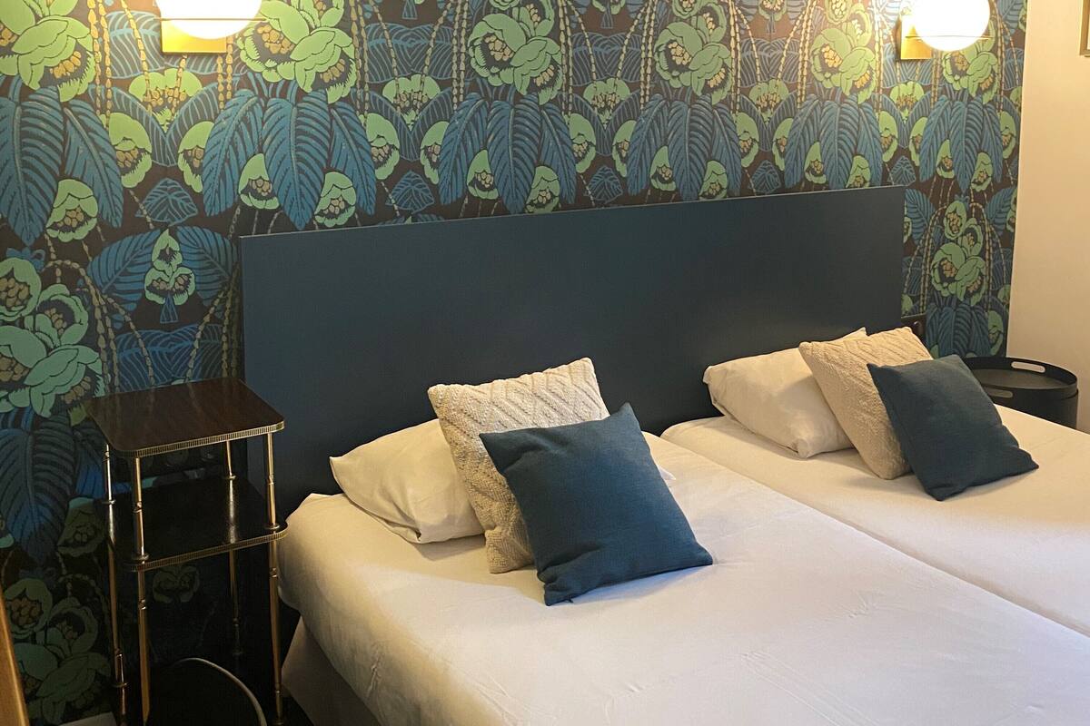 A hotel room with a patterned wallpaper, two bedside tables, a bed with pillows, and wall-mounted lights.