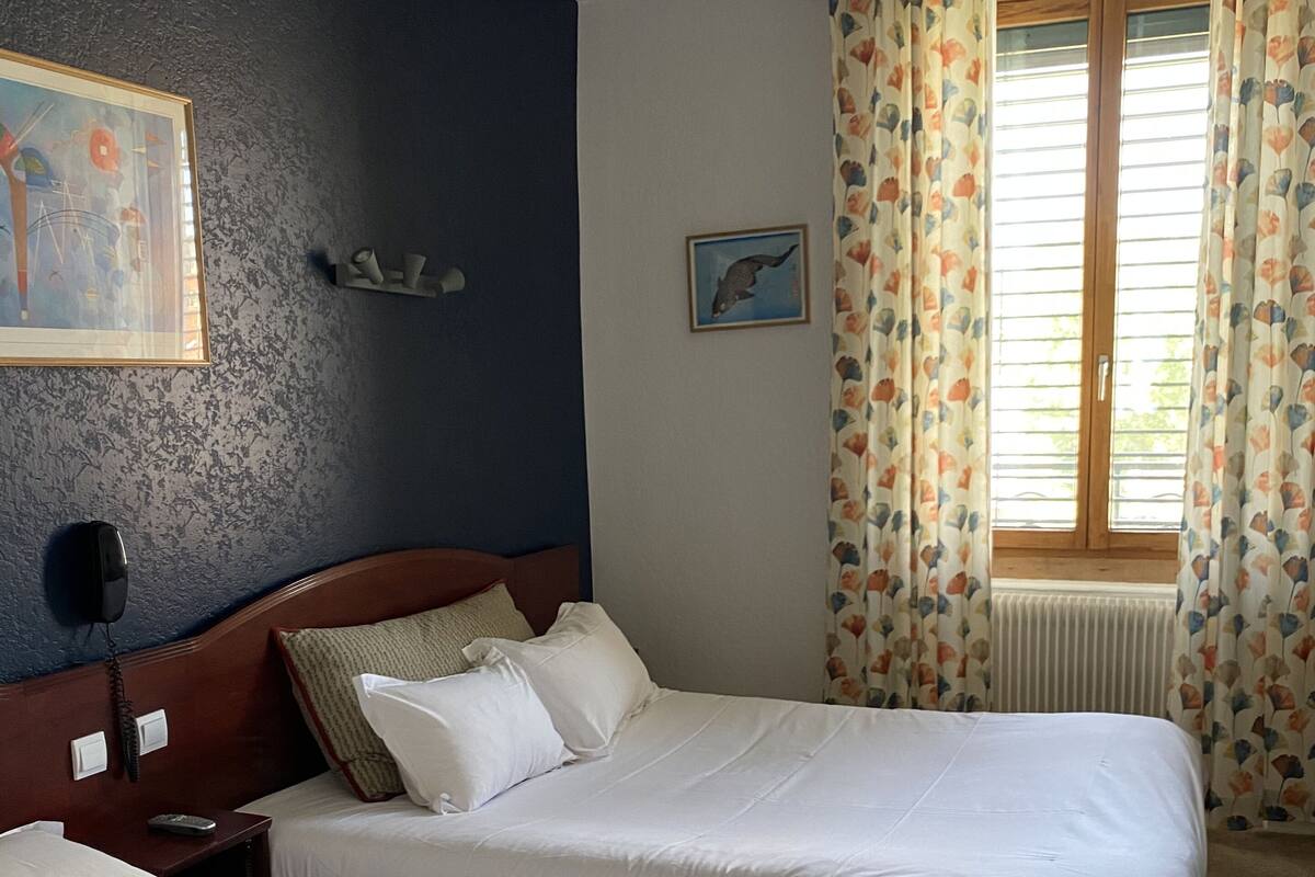 A hotel room with two beds, a headboard, a painting, a telephone, and a window with curtains.