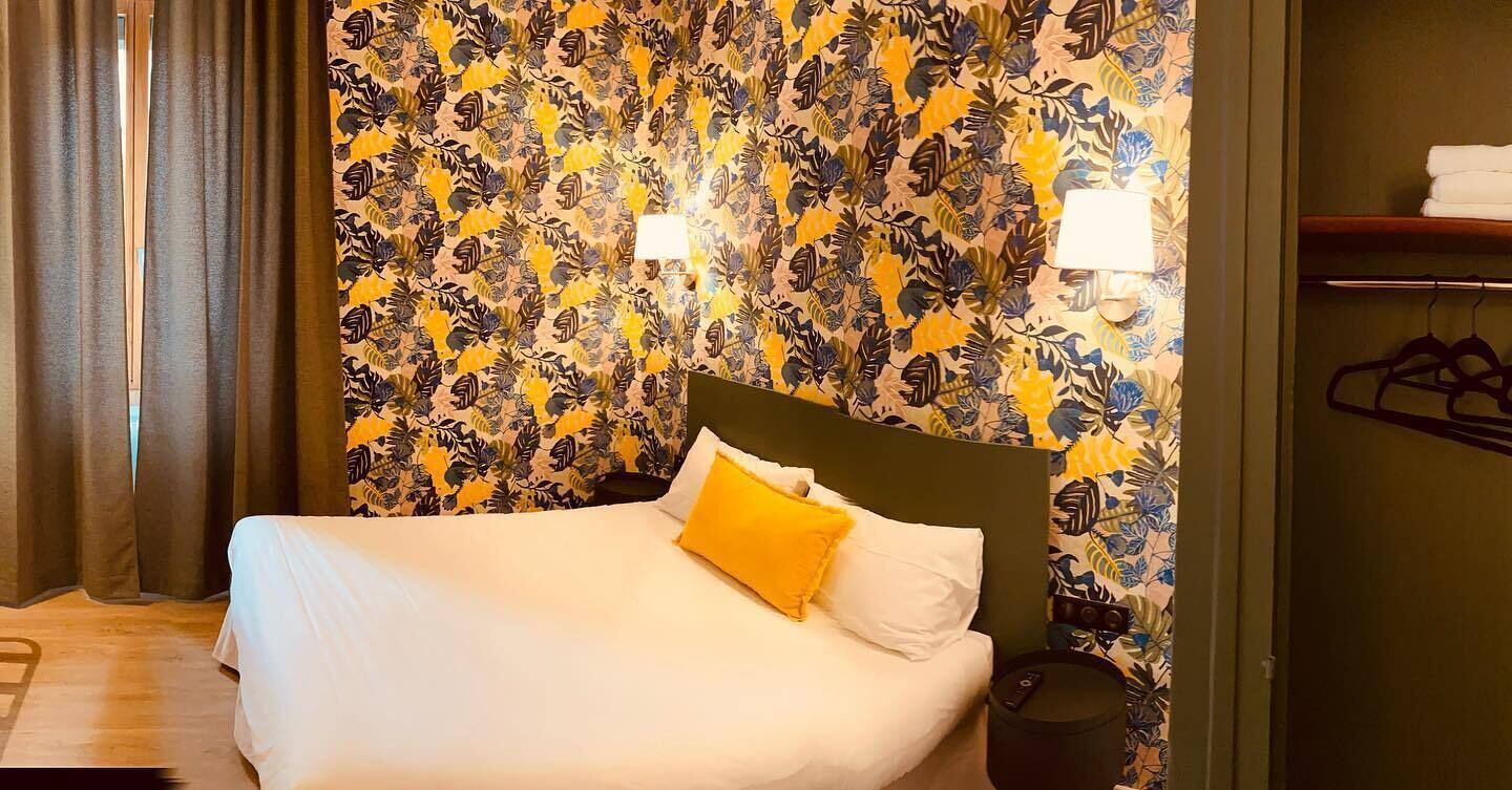 A hotel room with a bed, a headboard, a nightstand, a lamp, and a wall with a leaf pattern.