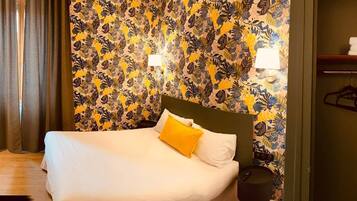 A hotel room with a bed, a headboard, a nightstand, a lamp, and a wall with a leaf pattern.