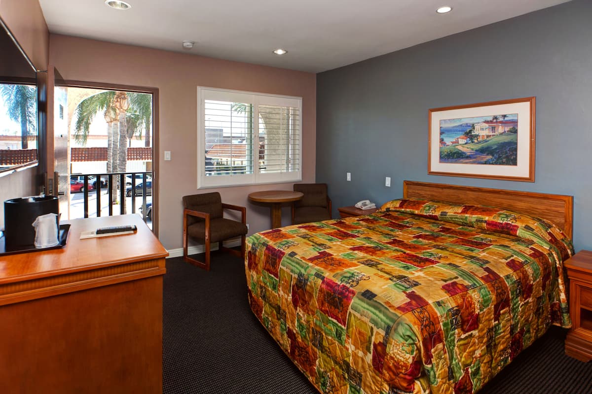 A hotel room with a large bed, a desk with a coffee maker, a chair, a small table, and a window with blinds.
