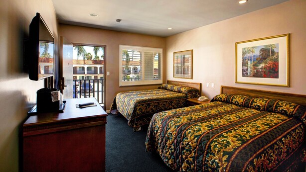 A hotel room with two beds, a desk, a chair, a television, and a window with a view of palm trees and buildings.