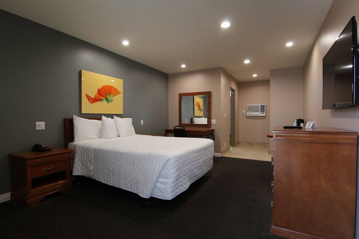 A hotel room with a large bed, a wooden nightstand, a desk, a mirror, and a flat-screen TV mounted on the wall.